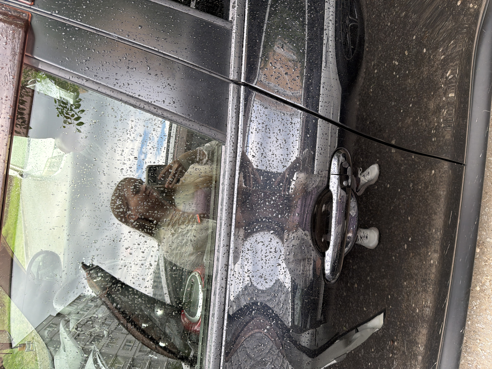 A person is reflected in the wet window of a car, holding a phone. The person is wearing a light-colored top and white sneakers. The car is dark-colored and appears to be parked on a street in Khabarovsk, Russia, as indicated by a license plate that is visible in the reflection and reads "982 XX 27". Water droplets are visible on the car's window and body, suggesting it is raining or has recently rained. The background shows buildings and greenery, typical of an urban environment. The image is taken from a low angle, looking up at the car window.