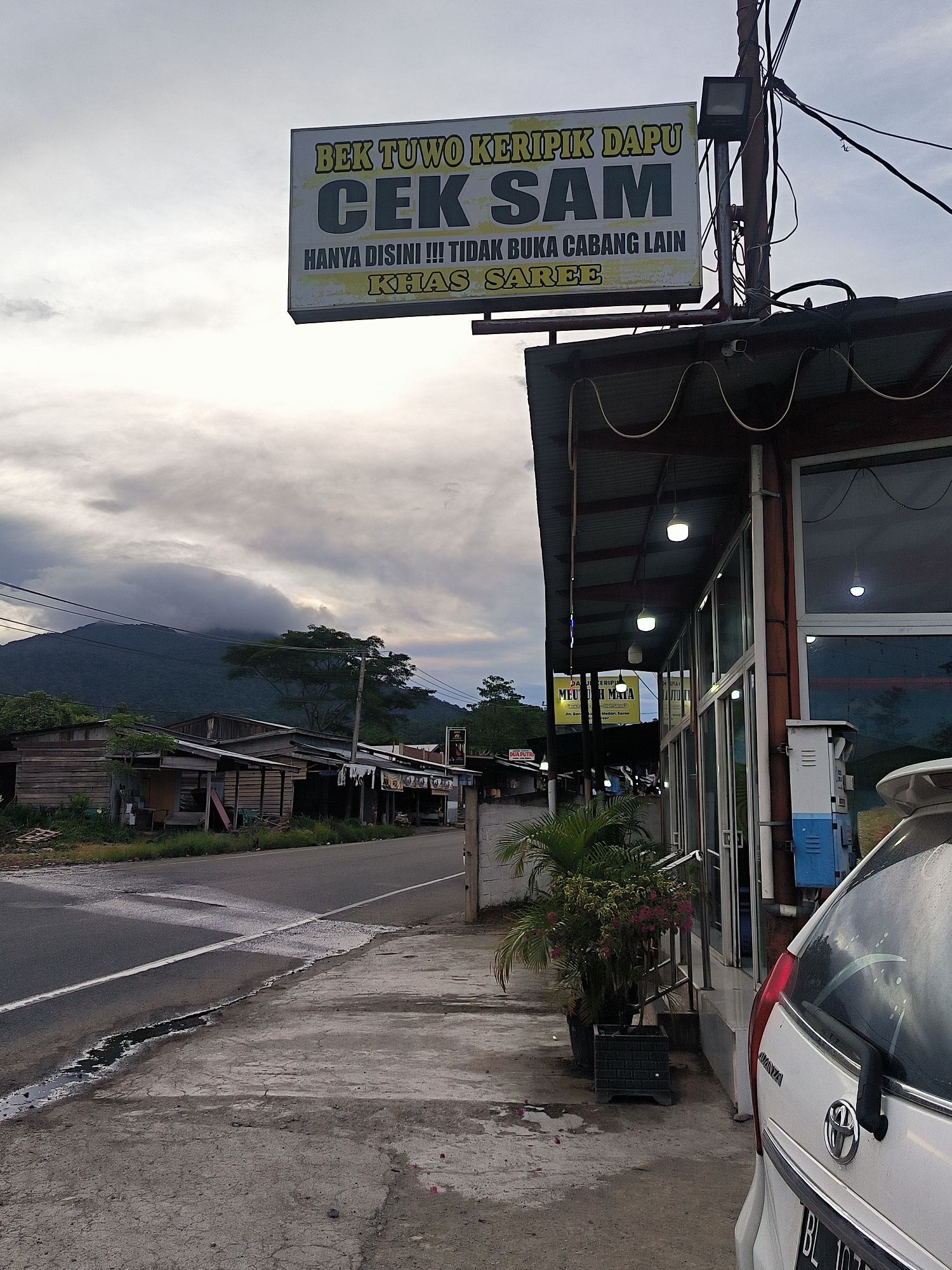 The image presents a quiet roadside scene in what appears to be a rural or semi-urban area, likely Sukamulia, Indonesia. The sky is heavily overcast, suggesting either late afternoon or early evening, with artificial lights already active on the building to the right.
A prominent rectangular sign hangs above this building, displaying text in Indonesian: "BEK TUWO KERIPIK DAPU CEK SAM." Below this, it reads, "HANYA DISINI !!! TIDAK BUKA CABANG LAIN" (Only here!!! No other branches opened) and "KHAS SAREE" (Saree specialty), indicating a business that specializes in crisps or chips. The building itself is a single-story structure with large windows, possibly a shop or restaurant, and has several pendant lights illuminating the area under its awning. A white car, partially visible, is parked in front of the building.
In the foreground, a damp asphalt road with a white dividing line runs alongside a concrete sidewalk. Across the road, a row of simple wooden shacks or houses can be seen. The background is dominated by a large mountain, its upper slopes and peak obscured by thick, low-hanging clouds, contributing to an atmospheric and somewhat muted scene. Trees are visible on the mountain and in the midground. Smaller, partially visible signs on the building's facade also mention "DUA PUTR," "MEU H MATA," and place names like "Medon" and "Saree." No people are visible, lending a tranquil and still quality to the image.