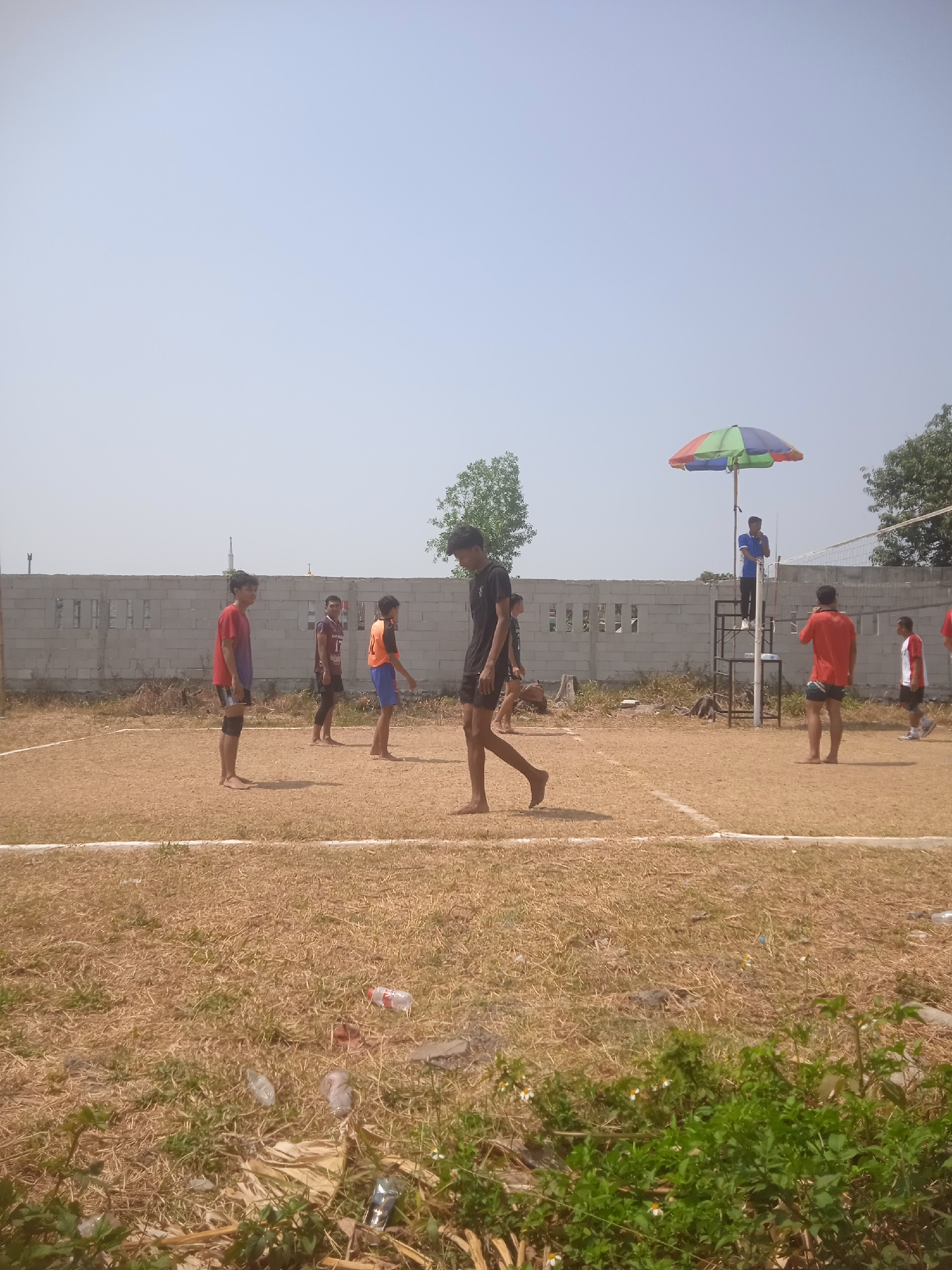 In Klodran, Indonesia, on a dry, grassy volleyball court marked with white lines, a volleyball game is underway. The sky is clear and pale blue. A light-colored brick wall runs along the back of the court. Several young men are participating in the game. One player, wearing a red shirt and black shorts, stands near the sideline, looking towards the net. Another player in a black shirt and shorts is in motion, possibly mid-play. A referee sits in a tall, elevated chair under a colorful umbrella, observing the game. Other players are positioned on the court, anticipating the next move. The environment is outdoors, with a few trees visible in the background. The ground is covered in dry grass and some scattered litter, including plastic bottles and wrappers. The scene captures a moment of athletic activity in a casual, outdoor setting.