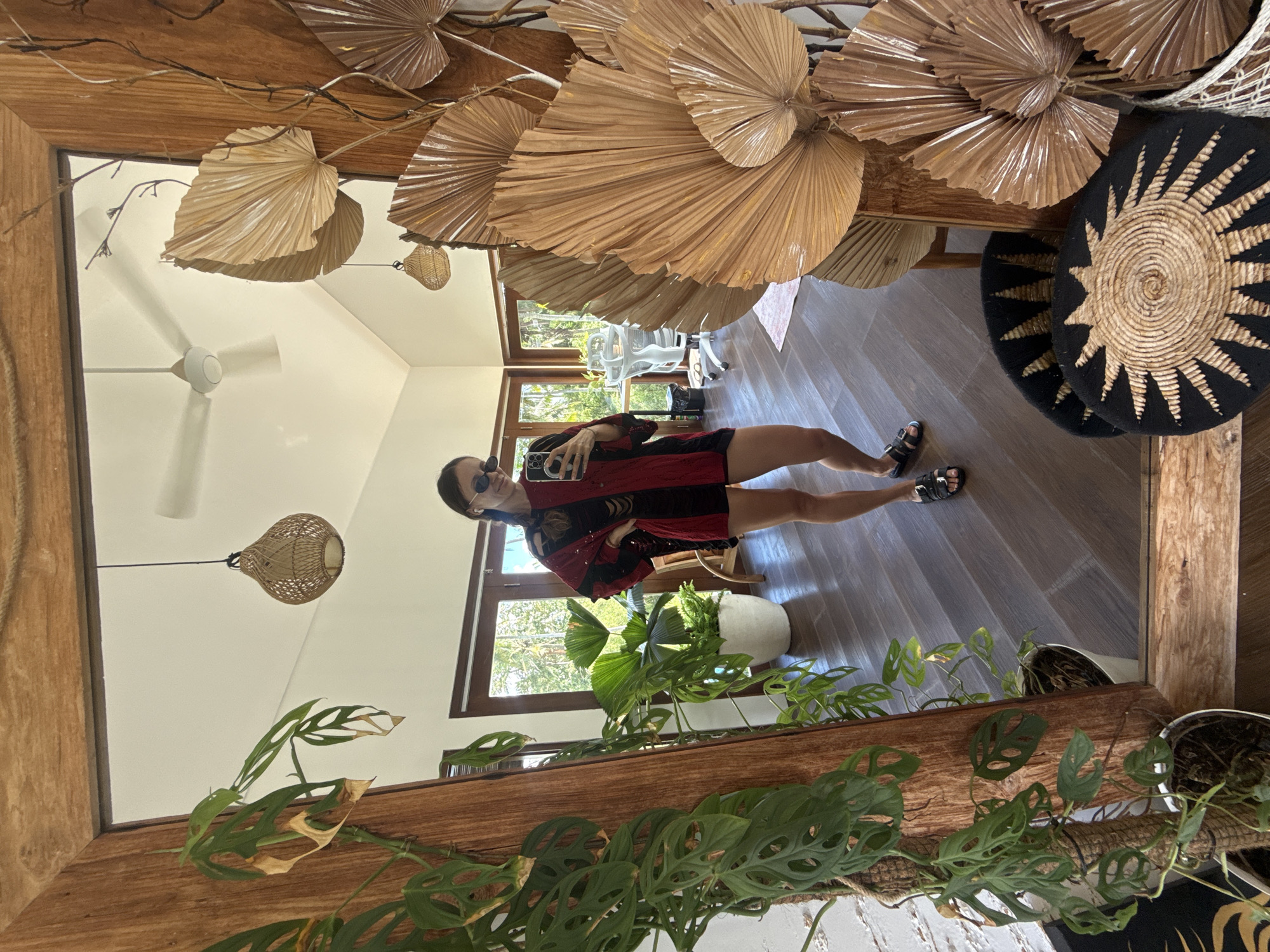 The image shows a reflection in a wooden-framed mirror. Reflected is a person wearing sunglasses, a red and black top, and short shorts. The person is holding a mobile phone, likely taking a selfie.
The reflection shows the interior of a room with a wooden floor. There is a ceiling fan. A variety of plants in pots are also reflected. Dried palm fronds are arranged above in an artful display. Woven wall hangings can also be seen.
Based on the decor and plant life visible, the image is likely taken in Mas, Indonesia, as indicated in the prompt.