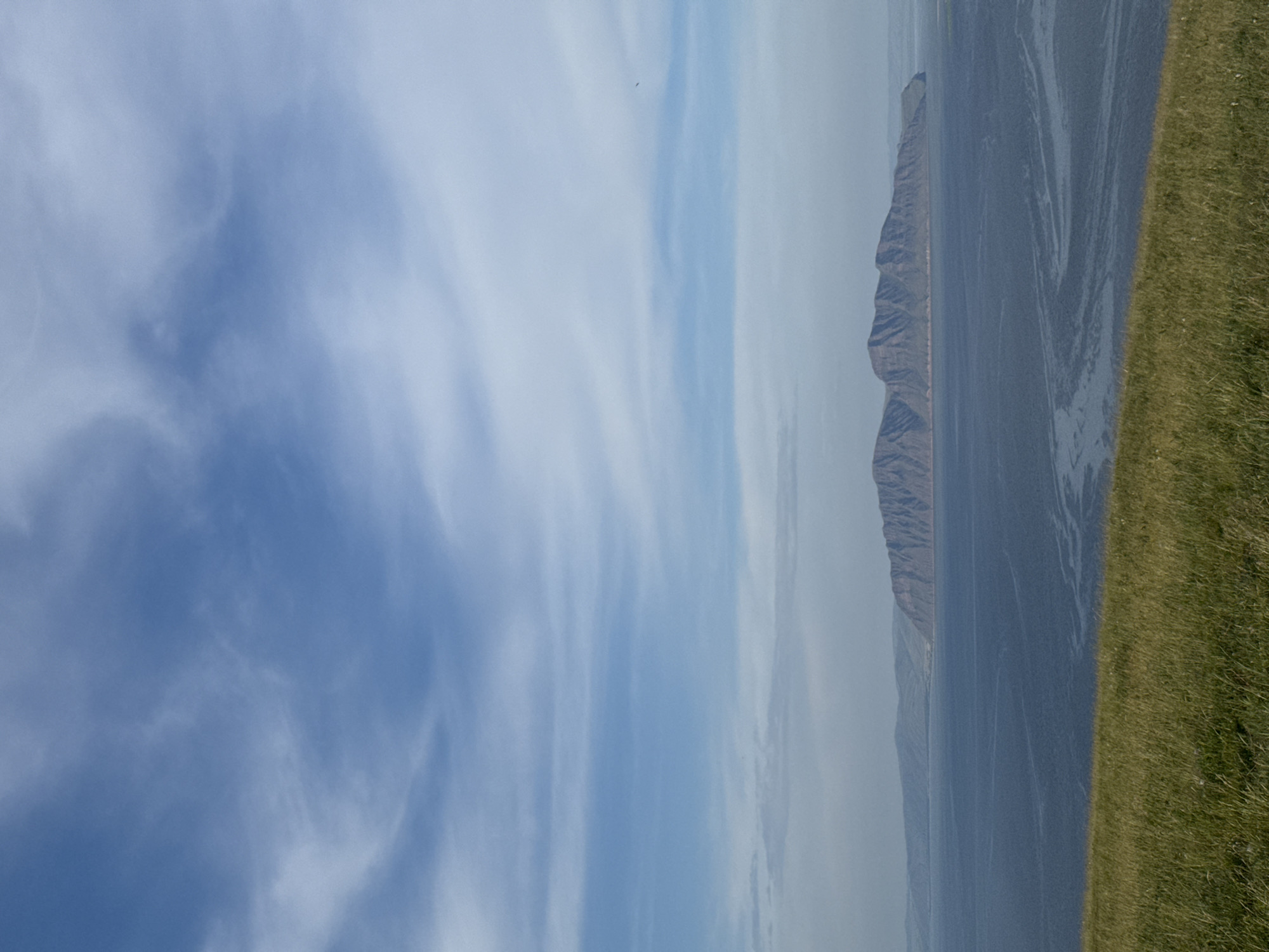 The image depicts a vast expanse of water, likely a large river or a reservoir, under a partly cloudy blue sky. In the distance, a long, rugged mountain range stretches across the horizon. The foreground is dominated by a grassy slope, suggesting a high vantage point overlooking the water and mountains. The scene is natural and serene, with no human presence or man-made structures visible. The lighting suggests it is daytime, possibly late afternoon or early morning, given the soft light. The location is identified as the city of Ust'-Abakan, Russia, implying this is a view of the Yenisei River or a related body of water in that region.