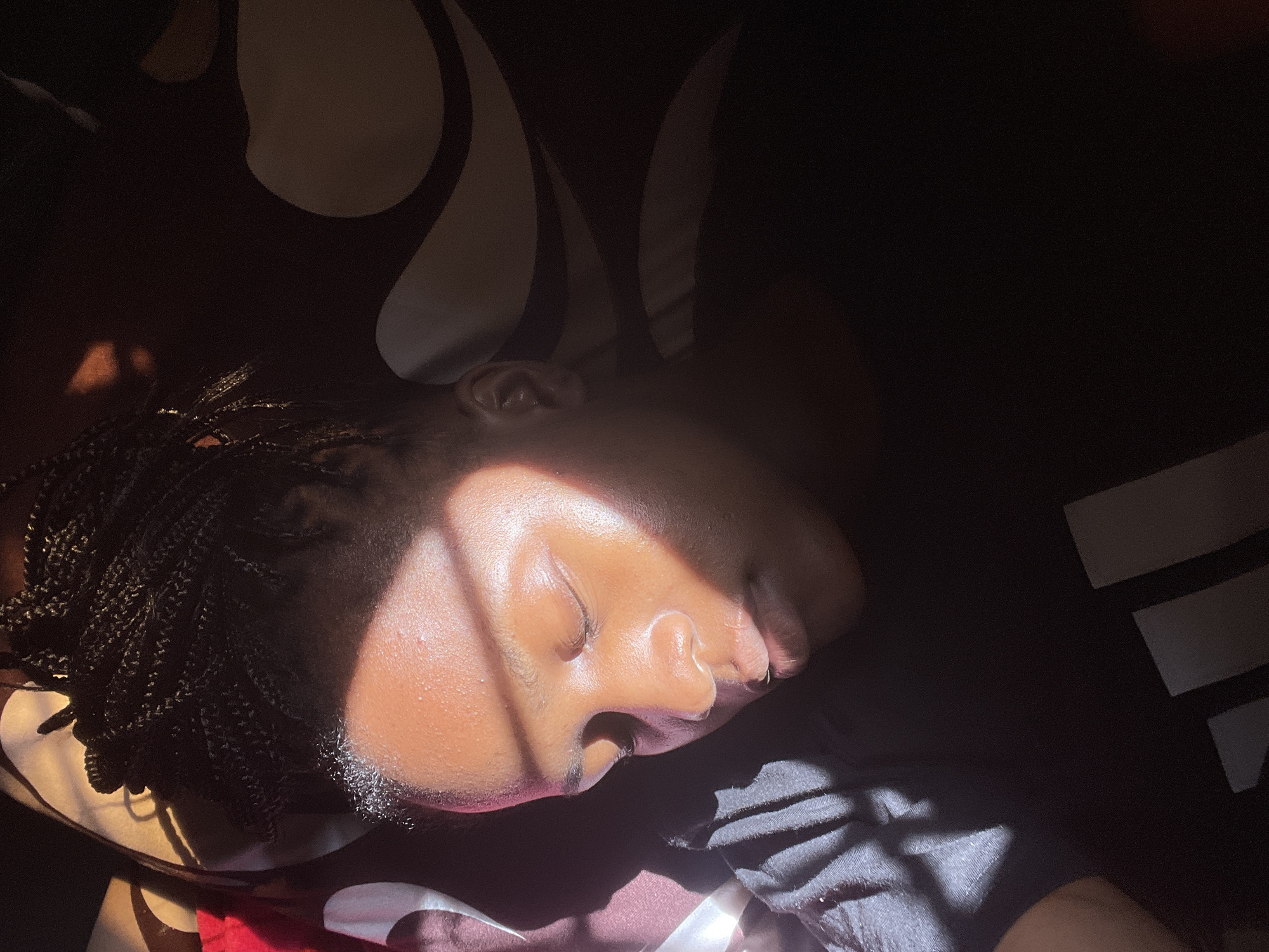 A young Black woman with braided hair is sleeping in a dark room, illuminated by a diagonal beam of sunlight across her face. She is lying on her side, wearing a black t-shirt with a white logo that appears to be three vertical bars. The background is a dark patterned fabric, possibly a bedspread or curtains. The light casts shadows across her features and the surrounding environment. The scene suggests a domestic setting within a residential area of Rafin Par, Nigeria.