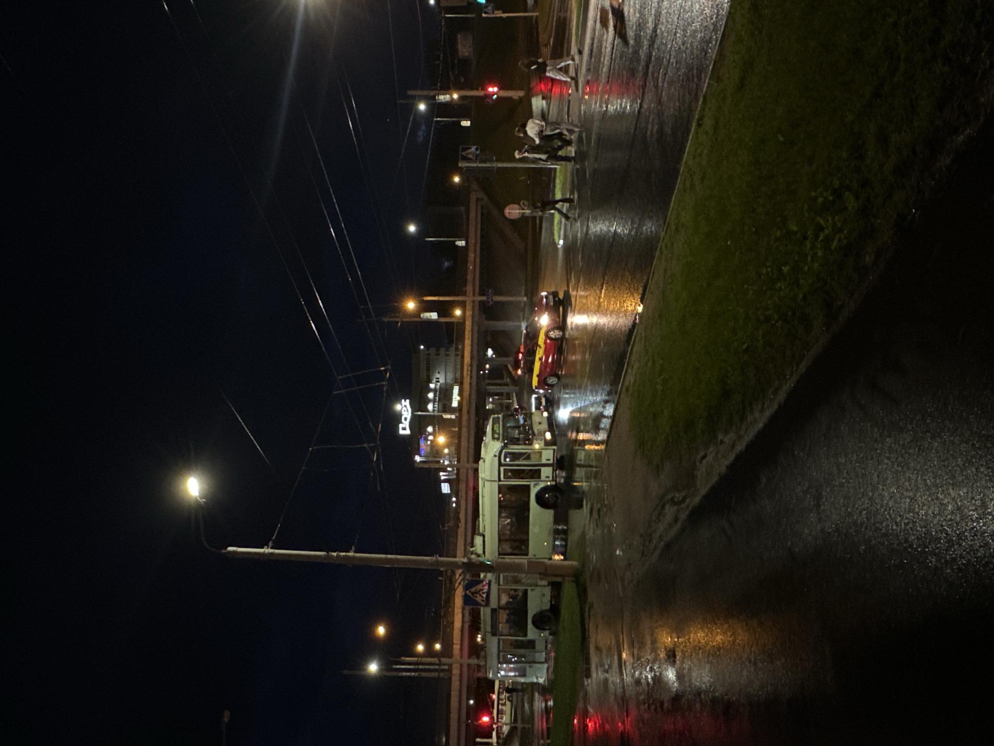 The image depicts a nighttime urban scene in Minsk, Belarus, characterized by a wet, reflective street surface. A trolleybus stands near a bus stop, likely awaiting passengers. Pedestrians are crossing at a crosswalk. There are streetlights that illuminate the area, and electric cables are visible overhead. The scene also includes cars and a grassy verge along the street. The wet surfaces reflect the city lights, creating a shimmering effect.