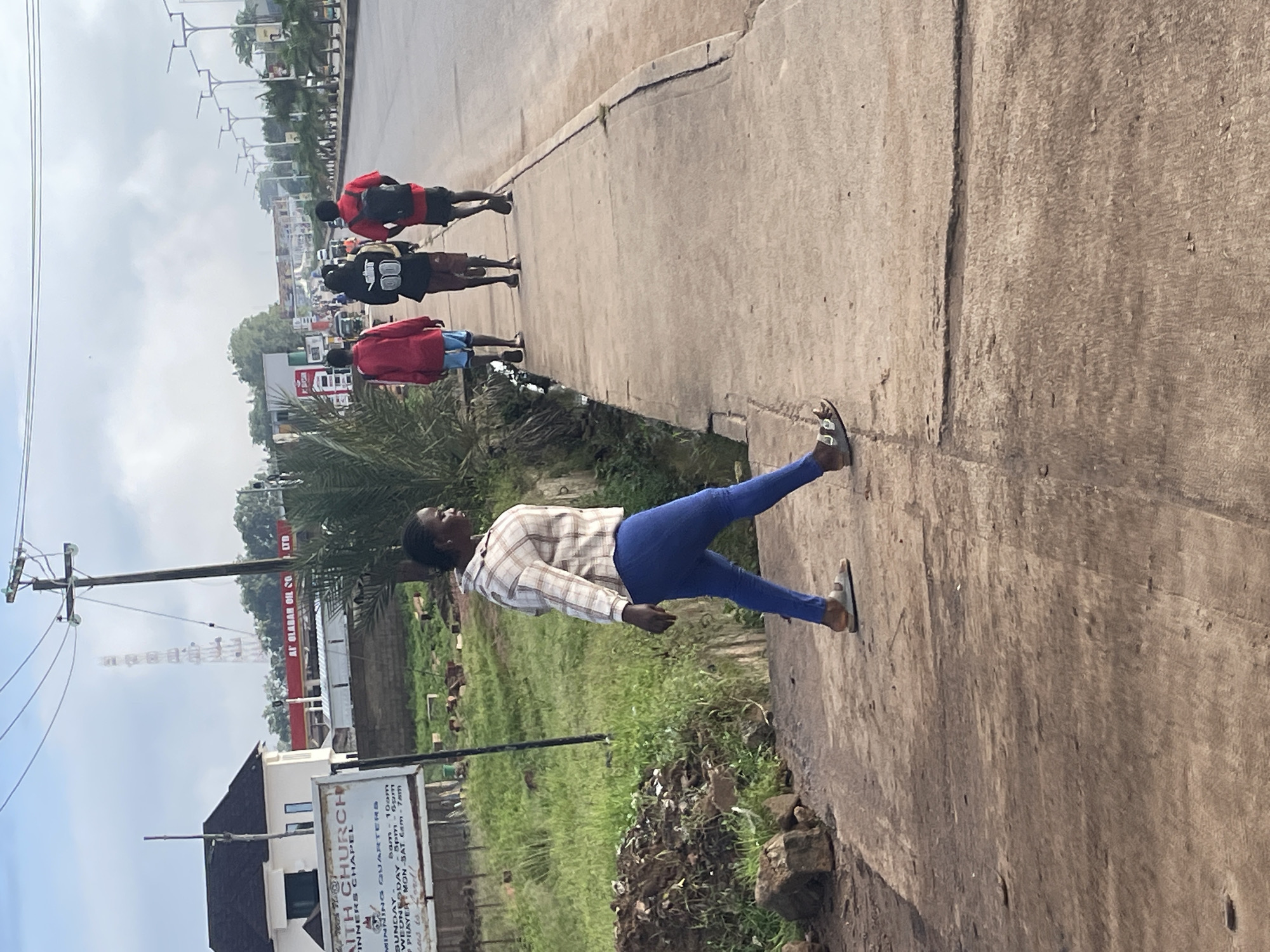 The image depicts a scene on a paved sidewalk beside a road in Jos, Nigeria, under an overcast sky. In the foreground, a woman wearing a checkered long-sleeved shirt and blue jeans is walking away from the camera, with her body angled to the right. She has dark hair pulled back and is wearing sandals. To her right, further down the sidewalk and walking away from the camera, are three young men. The one on the left wears a red jacket and light blue shorts, the middle one wears a black hooded jacket and dark shorts, and the one on the right wears a red shirt and dark shorts, carrying a black backpack. The sidewalk is cracked and worn. Beyond the sidewalk, there is a grassy area with some scattered debris and rocks. In the background, there is a white building with a dark roof, a sign that reads "FAITH CHURCH" and lists service times, and a gas station with a red awning labeled "OLABAN OIL SO LTD". Further in the distance, a telecommunications tower is visible against the sky, and the road appears to be a busy thoroughfare with some traffic and streetlights lining the right side. The overall impression is an urban street scene in Jos.