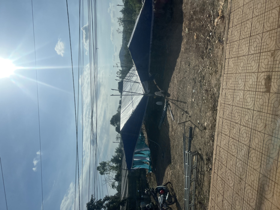 The image depicts an outdoor scene in Xuân Long, Vietnam, under a bright blue sky with scattered clouds and intense sun glare. Numerous black utility lines traverse the upper frame. In the foreground, a patterned light-brown concrete sidewalk is visible. Beyond it lies an unpaved, brown dirt lot containing two large, temporary canopy structures, featuring dark blue tarp roofs and white translucent sections, appearing to be in a state of assembly or disassembly. Light blue and white fabric hangs from the side of one canopy. Several metal support poles are evident, and multiple sections of metal scaffolding are scattered on the ground in the lower-left. At least two individuals, wearing light-colored clothing, are present near the canopies, engaged in activity related to the structures. A white scooter with red accents is parked on the left side of the dirt lot, near a dark fence. Green trees and distant hills are visible in the background.