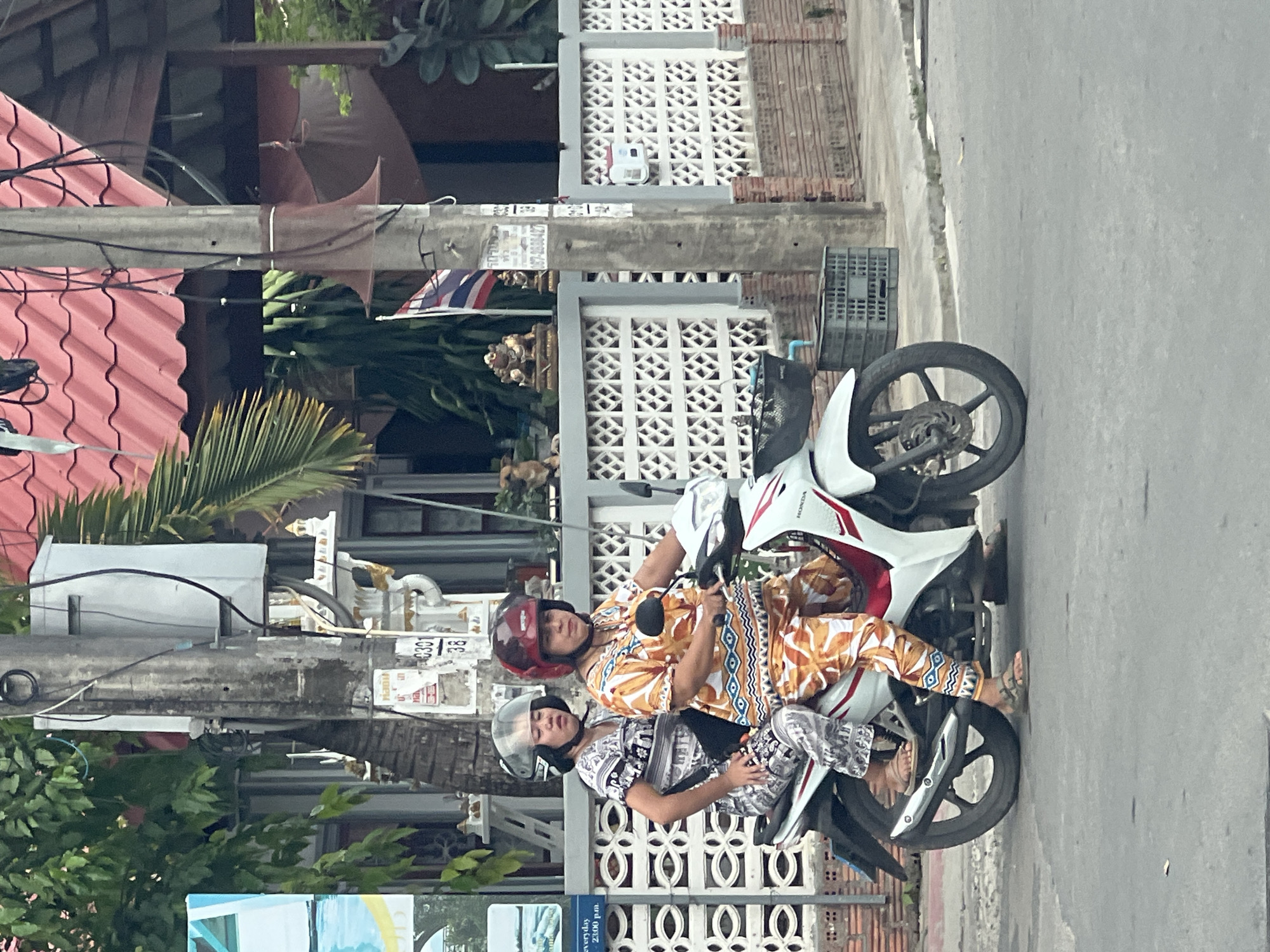 In บ้านบางรักษ์, Thailand, two individuals are riding on a white Honda motorcycle. The rider, positioned in front, wears an orange and white patterned shirt and pants, along with a maroon helmet. The passenger, seated behind, is dressed in a black and white patterned outfit and a clear helmet. They are traveling on a paved road with a grey asphalt surface, bordered by a sidewalk. To the left of the road, a white decorative fence with an intricate pattern is visible. Behind the fence, a utility pole stands tall, adorned with various signs and cables. Further in the background, a building with a red tiled roof and palm trees are present. The scene captures a moment of everyday life on a street in this Thai locale.