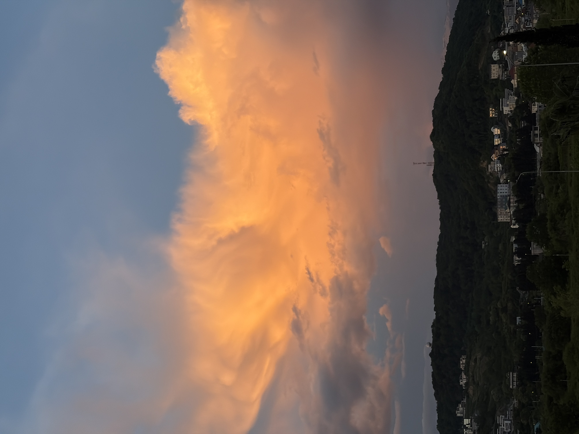 The image shows a view from the city of Sochi, Russia. The landscape is divided between the sky on the left and a dark, wooded hill on the right. The sky features large, peach-colored clouds that are illuminated by the setting sun. The clouds are dense and textured, with varying shades of orange and pink. To the right of the clouds, the sky darkens to a more muted, grayish-purple hue. The hill is covered in dense trees and houses, with some buildings showing light. There is a pole-like structure atop the hill. Street lights are visible on the hill. The overall atmosphere is serene and captures a moment of transition from day to night.
