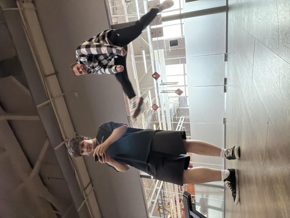This image depicts an indoor scene in Chaikovsky, Russia, likely within a modern commercial or recreational facility. The floor is composed of light-colored wood-effect laminate. Two individuals are present.
On the left, a young person with short, dark, curly hair stands facing the camera, holding a dark smartphone with both hands. They are wearing a black short-sleeved t-shirt with white Russian text ("ДЕЛА НЕТ"), black shorts, white socks, and black sneakers with white soles. A silver chain is visible around their neck. Their expression is a slight smile.
On the right, an adult male with short, dark hair is captured in mid-air, performing a dynamic jump or kick. He is wearing a black and white checkered long-sleeved shirt, black athletic pants with a white Nike swoosh on the left thigh, and light-colored athletic shoes. His right leg is extended horizontally to the left, and his left leg is bent. Both arms are bent at the elbows, with hands clenched into fists near his chest. His mouth is slightly open, conveying a sense of exertion or a posed expression.
The background features large glass partitions, some frosted in the lower half, through which another interior space is visible. A sign affixed to the glass reads "ВОЛЖСКИЙ КАФЕ" (Volzhsky Cafe) in Russian. Several red diamond-shaped signs with white pictograms and a horizontal rectangular sign are also visible on the glass. Overhead, the ceiling displays exposed light-colored beams and ductwork. A portion of a black exercise machine, possibly a treadmill, is visible on the far left. Natural light emanates from windows beyond the glass partitions.