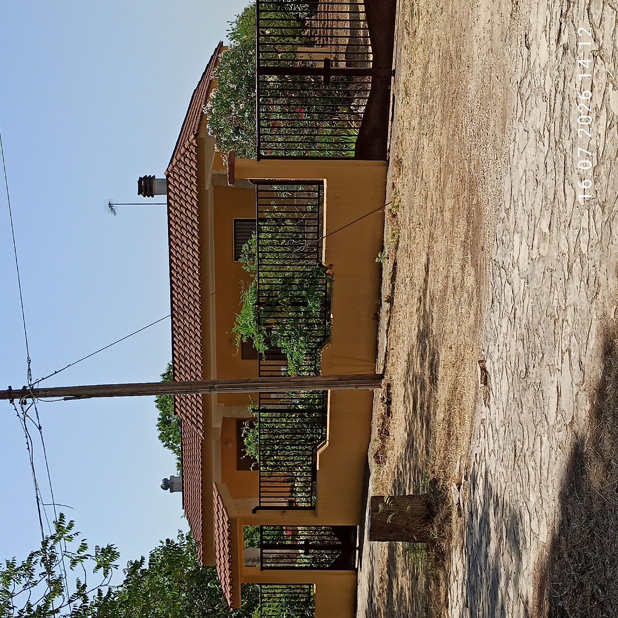 This is a medium-shot, eye-level photograph of a single-story house in Grumo Appula, Italy, on a clear, sunny day. The house has a yellow stucco exterior and a terracotta tile roof. A black metal fence encloses the property, with a black metal gate on the left side leading to the entrance. To the right, there is another black metal gate. The yard is dry and dusty, with patches of dry grass and a paved area in the foreground made of irregular, flat stones. A wooden pole with electrical wires runs vertically on the left side of the frame, and a second, thinner pole is visible on the right. There is a small tree stump in the foreground to the left. The image contains a timestamp in the lower right corner: "16.07.2026 14:12".