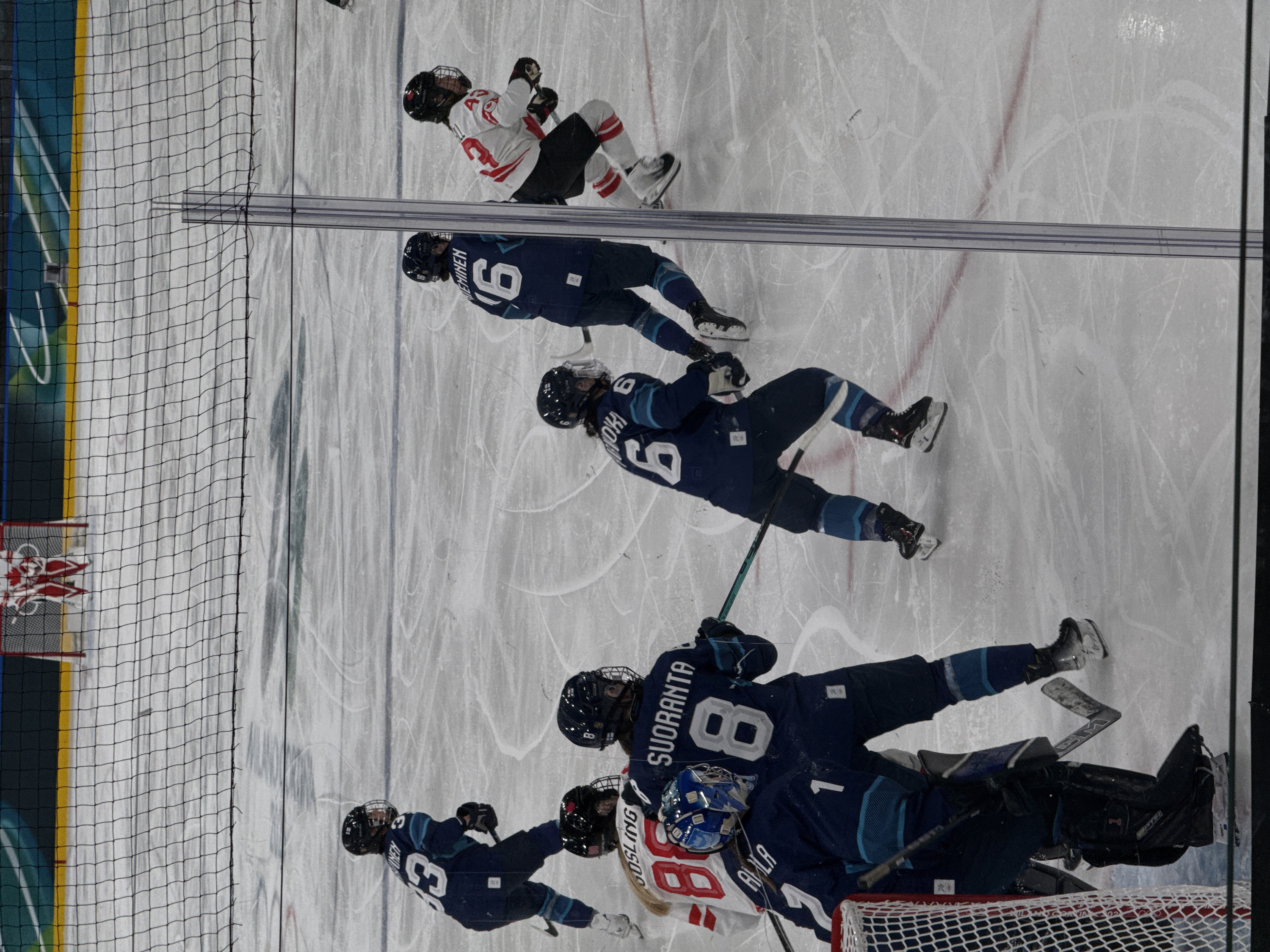 An ice hockey game is depicted on an ice rink in Rho, Italy, observed through a black protective net that occupies the foreground. Eight players are visible on the ice surface.
Five players wear dark blue jerseys with light blue chevron patterns and white numbering. This group includes a goaltender, identified by the number '1' on the leg pad and 'AMALA' on the blue helmet, positioned in front of a red-framed goal net in the lower left. Four additional skaters in dark blue jerseys are present: Player #8 with 'SUORANTA' on the back, standing near the goaltender; Player #3 (name partially obscured) in the upper left, facing right; Player #6 (name partially obscured) in the center-right, facing away; and Player #6 'HEMINEN' in the mid-right, facing left.
Three players wear white jerseys with red accents and numbering. These include Player #88 'GOSLING' (partially visible) positioned near blue team Player #8, and Player #43 (name obscured) in the mid-right, actively skating towards the left. A second goaltender, dressed in red and white equipment, is partially visible through the net in the distant background, in front of another red-framed goal.
All players are fully equipped with ice skates, helmets with face cages, and hockey sticks. The ice surface displays extensive skate marks. The rink's perimeter features a yellow kick plate beneath dark blue boards, which include white design elements. Vertical transparent barrier supports are visible in the foreground and mid-ground structures.