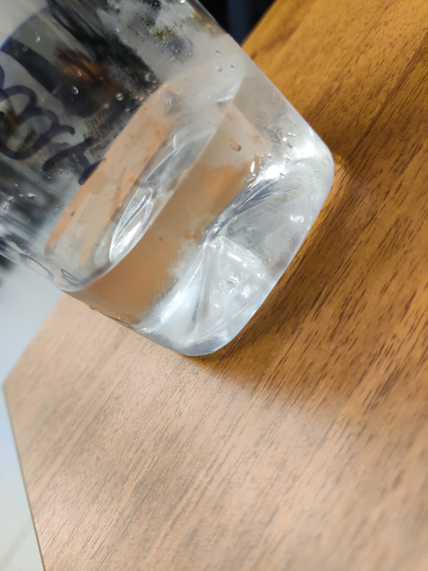 The image is a close-up, angled shot of a clear plastic bottle filled with a clear liquid, resting on a wooden surface. The bottle's bottom corner is visible, showing condensation on the outside. The liquid inside has some slight opaqueness, possibly due to dissolved substances or air bubbles. The wooden surface has a visible grain pattern and appears to be a tabletop. The background is out of focus, showing some dark and light shapes that are not clearly identifiable. There are no people or discernible actions taking place in the image. The image does not provide any information to determine its location in Neftekachka, Russia.