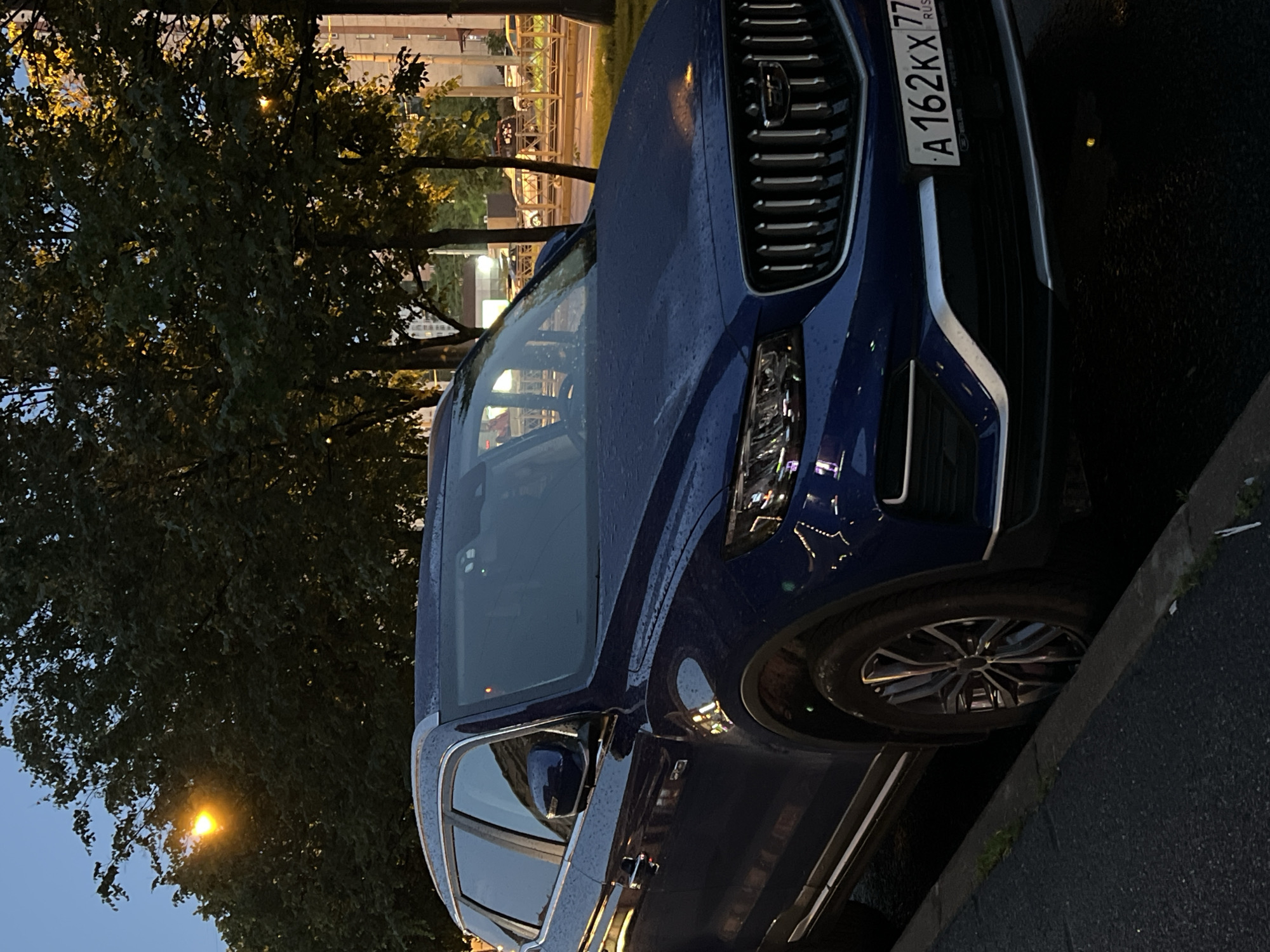 This media file shows the front passenger side of a dark blue car parked on a street. The car is wet, suggesting it might be raining or has recently rained. The time of day appears to be dusk or early evening, as indicated by the dim natural light and the illuminated street lamp visible in the background. Trees with dense foliage are also present, casting shadows and partially obscuring the background.
The car has a prominent chrome grille and a license plate with the characters "A 162KX 77 RUS". The headlights are visible, and the car's paint is reflecting the ambient light, creating highlights on its wet surface. The scene is set on a paved street, with a curb visible in the foreground. The overall mood is calm and subdued, typical of a quiet urban or suburban street in the evening. There are no people visible in the image. The location context is unknown, but the license plate suggests it is in Russia.