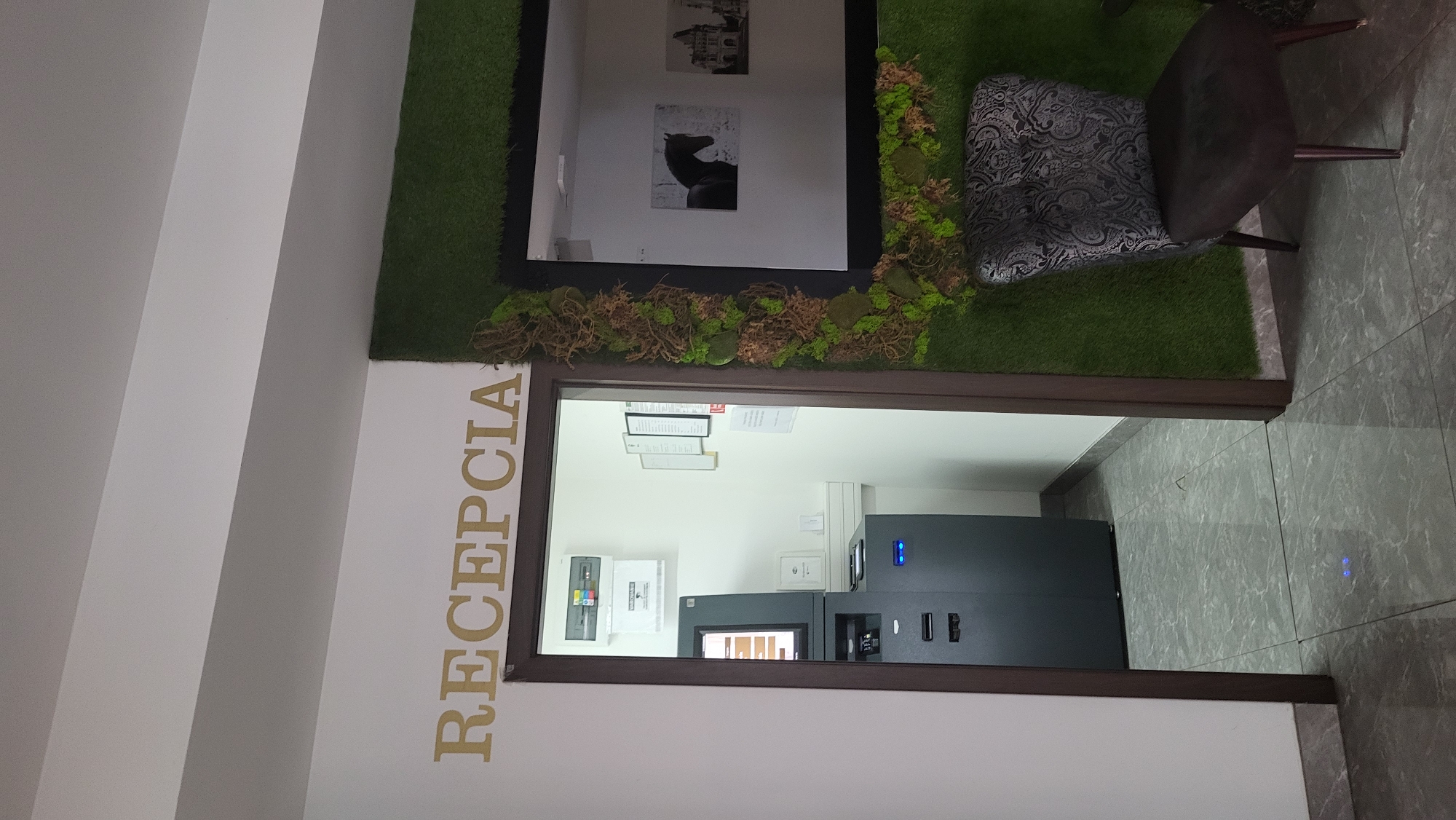 The image depicts an interior scene that appears to be a reception area. A doorway is visible, leading into a small room with an ATM machine and what looks like a distribution box on the wall. Above the doorway, the word "RECEPCIA" is spelled out in gold lettering. To the right of the doorway, a wall is covered in artificial greenery and moss, with a framed mirror mounted on it. Within the mirror, a black and white image of a horse is visible, along with another architectural image. In front of the green wall, a dark upholstered armchair with an ornate pattern is placed. The floor is tiled with a grey marble-like pattern. The overall impression is a modern and somewhat stylized reception area. The location of this scene in the city of Prešov, Slovakia, is unknown based solely on the visual information provided in the image.