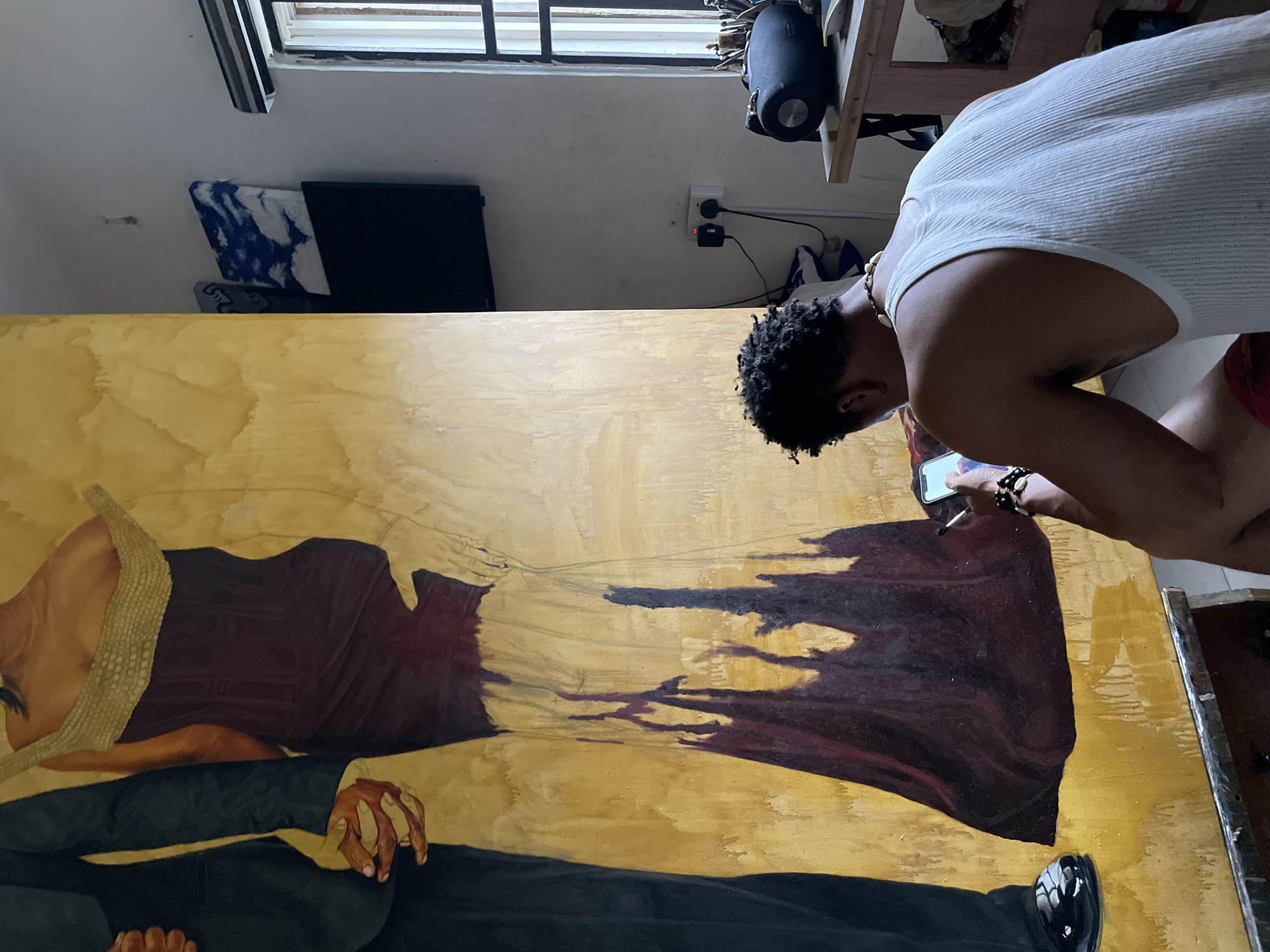 In Ado, Nigeria, an artist, seen from behind, works on a large canvas in a studio. The canvas depicts two figures, a man in a dark suit and a woman in a deep red dress with a textured gold top. The artist, wearing a white ribbed tank top and a beaded necklace, holds a smartphone and a paintbrush, appearing to reference the phone while painting. The studio environment includes a window with blinds, a shelf with a speaker and art supplies, and electrical outlets on the wall. The overall lighting suggests a daytime setting.