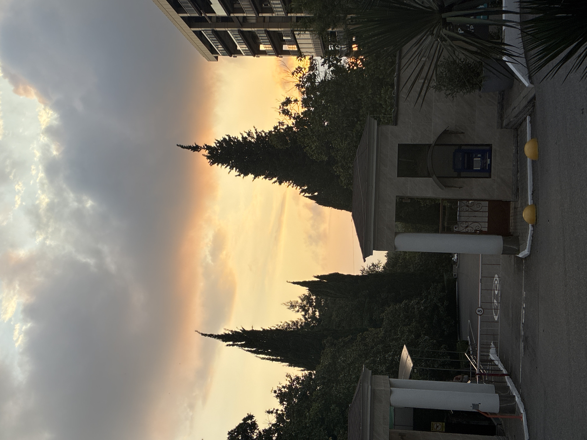 The image depicts a view of an entrance gate and a modern building against a sunset sky. The gate is partially open and equipped with a barrier. To the left of the gate, a white structure with columns is visible. To the right, a multi-story building with balconies is prominent. Several tall, slender cypress trees are silhouetted against the colorful sky, which displays hues of orange and yellow with clouds. A blue ATM machine is mounted on the wall of a small building near the gate. Yellow bollards are placed in front of the gate. The foreground shows an asphalt road. The scene appears to be a residential or hotel complex, possibly located in Sochi, Russia, given the architectural style and vegetation common to the region.