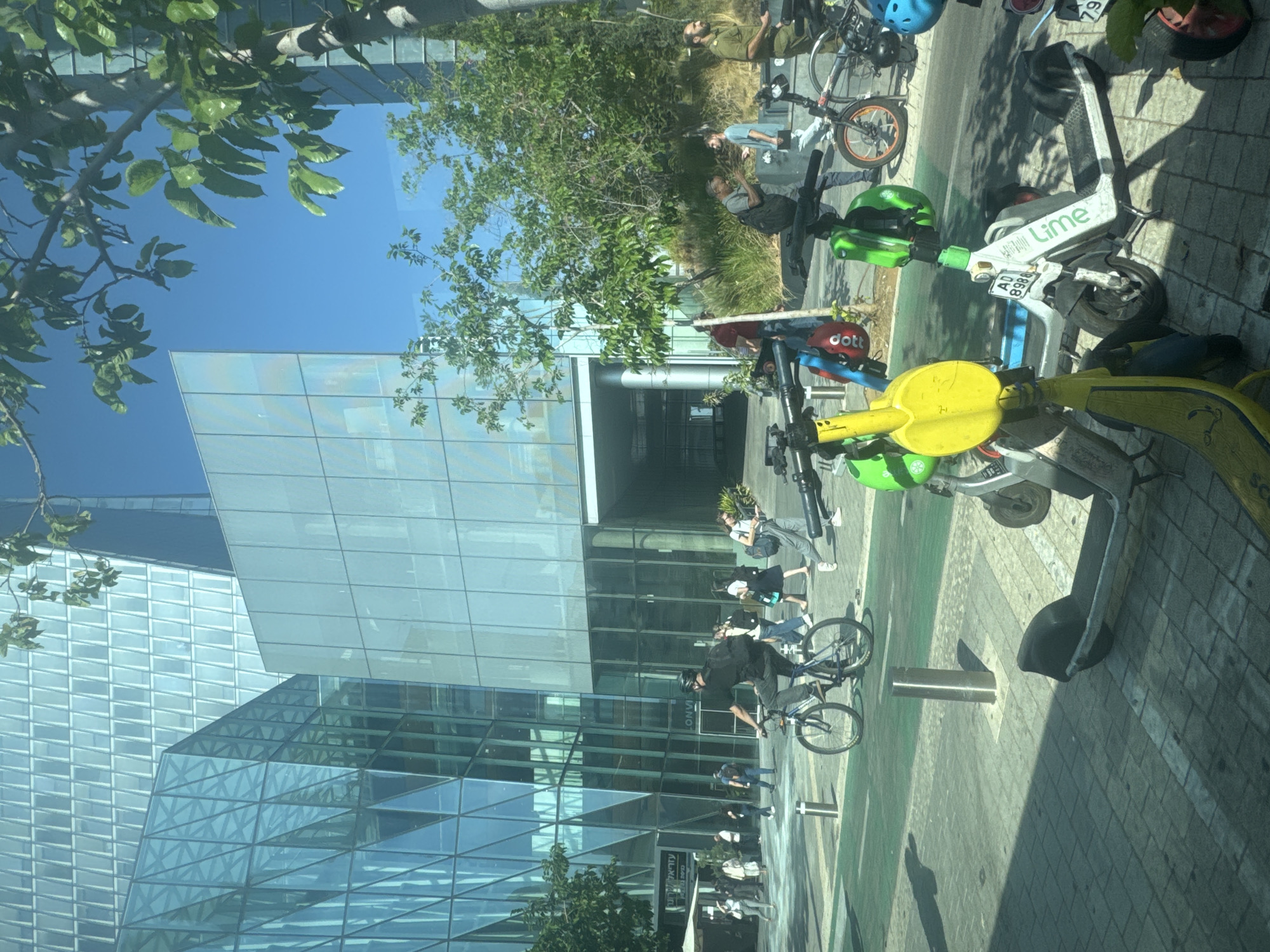 The image captures a sunny day scene in Tel-Aviv, Israel, likely on a pedestrian or bike path in front of a modern glass-facade building. In the foreground, several electric scooters are parked, two prominently displaying the "Lime" brand. One yellow scooter is positioned to the left, and a white and green Lime scooter with license plate "AD 8988" is in the center. To the right, another scooter with a blue helmet is partially visible.
In the middle ground, a person on a bicycle with a black backpack rides on a green-striped lane. Behind the cyclist, a group of pedestrians are walking and standing in front of the building's entrance. Some of these individuals are carrying bags and wearing casual attire. The building itself features an angular, modern design with extensive glass panels that reflect the bright sky and surrounding greenery.
Further back, on the right side of the image, a man in a military-style uniform stands. Other individuals are visible near the building entrance and among the trees. The environment is urban, with paved walkways, street furniture like bollards, and trees providing shade. The sky is clear and blue, indicating pleasant weather.