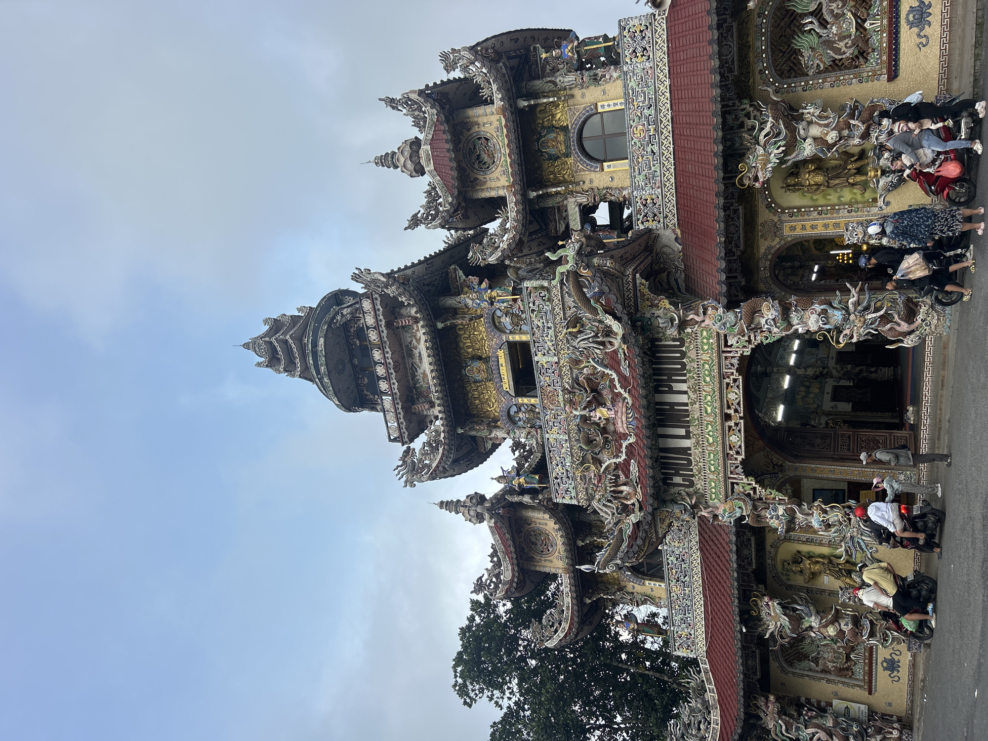 The image shows the entrance to the Linh Phuoc Pagoda in Xuan Thanh, Vietnam. The pagoda's facade is intricately decorated with mosaics and sculptures, featuring dragons and religious figures. Several people on motorbikes are parked along the side of the street in front of the pagoda.