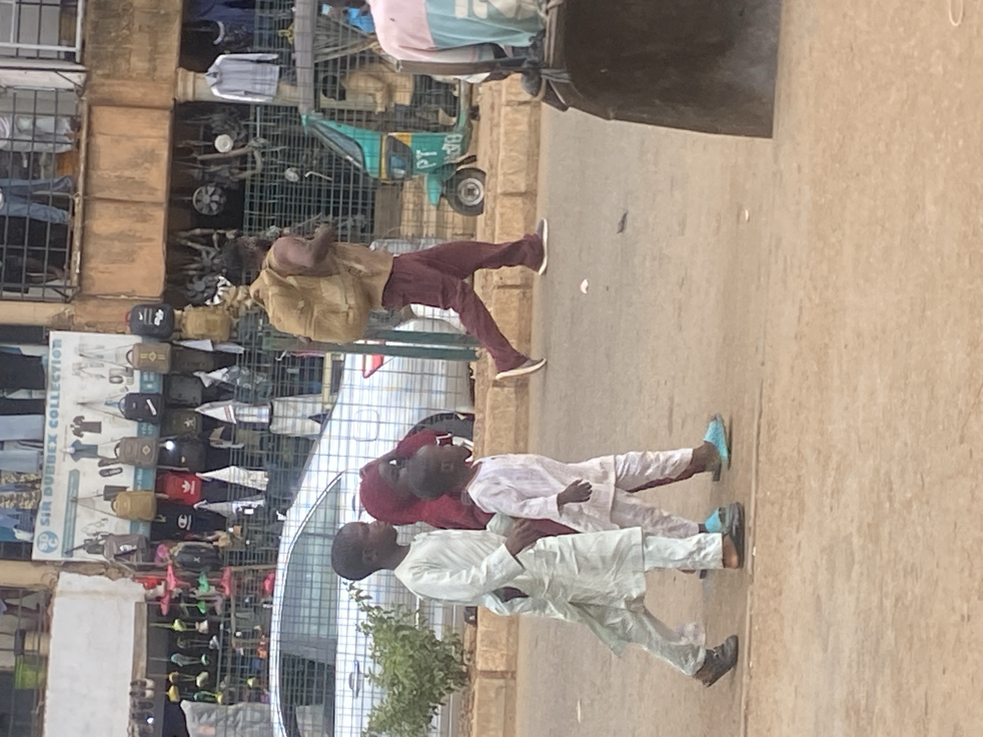 In Bukuru, Nigeria, a street scene unfolds with a marketplace and pedestrians. A man wearing a tan backpack and maroon pants walks on the right side of the road. To his left, three children are walking. The oldest, a boy in a white outfit, is on the far left. Next to him is a girl in a red head covering and dark dress, and a younger boy in a white outfit is on the far right of the group. Behind them, a silver car is parked. In the background, a building with shops displaying clothing and bags is visible. A green and yellow auto-rickshaw is parked to the right of the car. The ground is paved with a reddish-brown material, and a low concrete barrier runs along the sidewalk. The overall impression is of a busy commercial area.