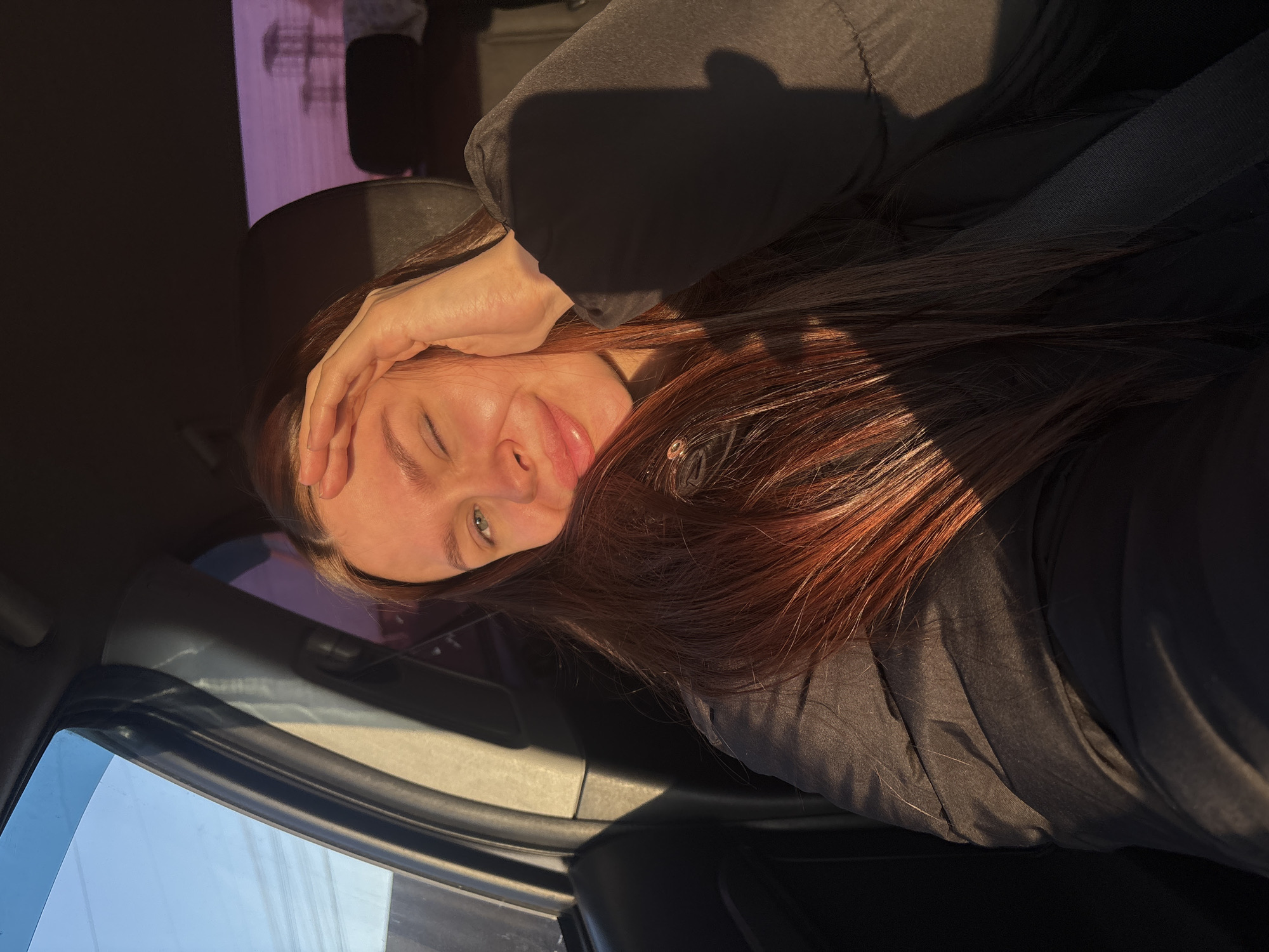 An individual is depicted seated within the interior of a vehicle, identified as being in Chaikovsky, Russia. The subject is a young adult female with long, reddish-brown hair, illuminated by direct sunlight from the right, casting shadows on the left side of her face and body. She is winking her right eye, smiling, and her left hand is positioned on her forehead. She wears a dark, possibly padded jacket, and a seatbelt is visible across her torso. The car's interior is dark. Through the front passenger window, a clear blue sky is visible. Through the rear window, a muted purple-hued sky or reflection is seen, with indistinct structures, possibly power line towers, in the distant background.