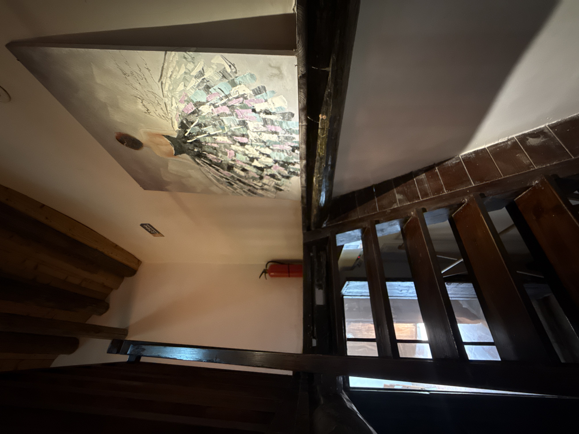 The image shows an interior staircase with wooden steps and dark wooden railings. On the wall above the staircase, there is a painting of a ballerina in a dark dress with a full, colorful skirt. To the left of the painting, there is a red fire extinguisher mounted on the wall. The ceiling consists of exposed wooden beams. The scene appears to be in a building, possibly a home or a small establishment. There are no people visible in the image. The lighting suggests an indoor environment, possibly with natural light coming from an unseen window. The location is identified as Longgang District, China.
