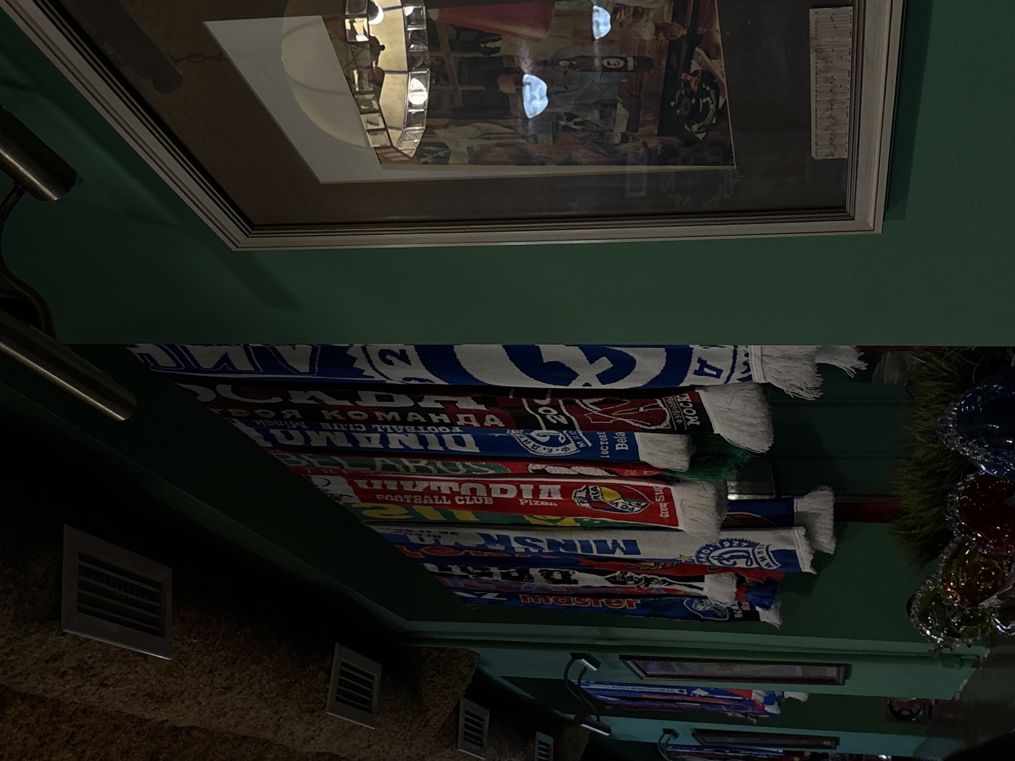 The image shows a collection of football scarves hanging on a wall, likely within a sports bar or a similar establishment in Minsk, Belarus. The scarves are stacked one on top of the other, displaying the names of various football clubs and their respective city locations, including "Minsk," "BELARUS," "Pizen," and "MOCK." A framed picture hangs above the scarves. Ventilation grates and ceiling lights are also visible, along with decorative plant life and hanging items.