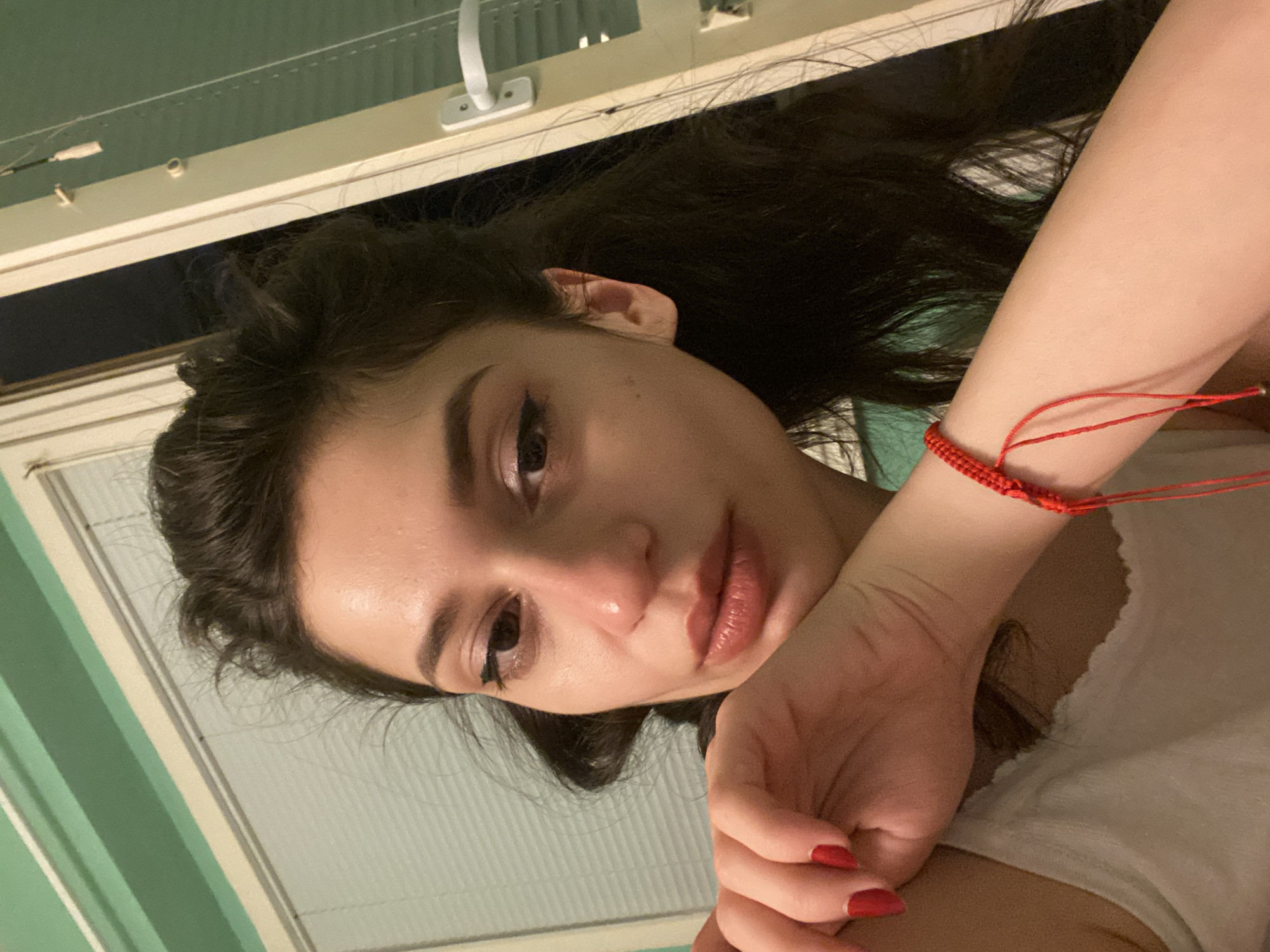 A close-up, medium-shot, eye-level, indoor selfie features a young woman with dark hair and makeup, her right hand resting on her chin. She wears a white tank top and a red bracelet on her left wrist. The background consists of a green wall and a window with blinds. No specific location in Sofia, Bulgaria is discernible from the image.