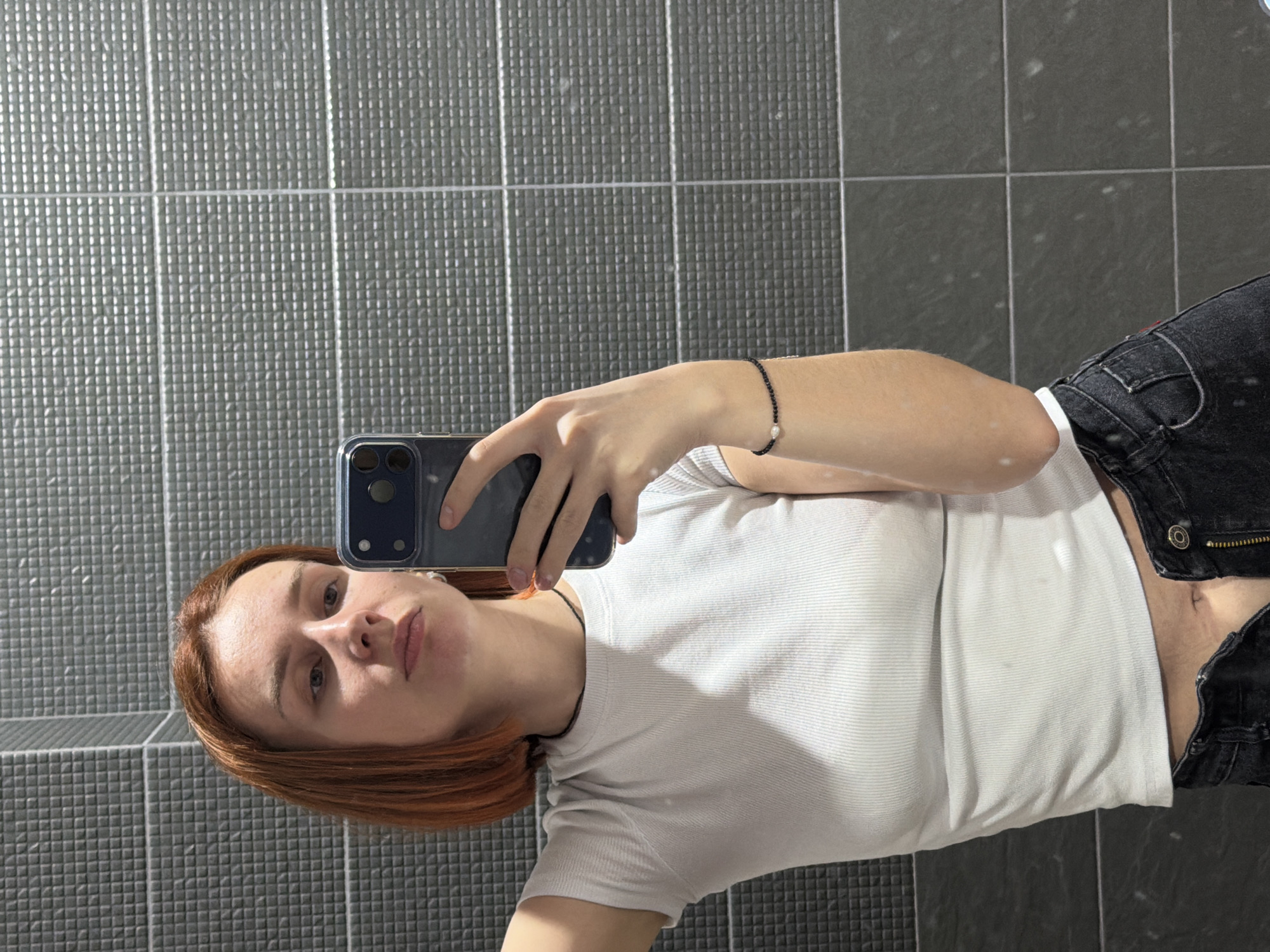 A young person with auburn hair is taking a selfie in a bathroom. The person is wearing a white t-shirt and black jeans. A black beaded bracelet with a pearl is visible on their right wrist. The bathroom has dark grey tiled walls. The location is unspecified; information about Chaikovsky, Russia is not discernible from the image.