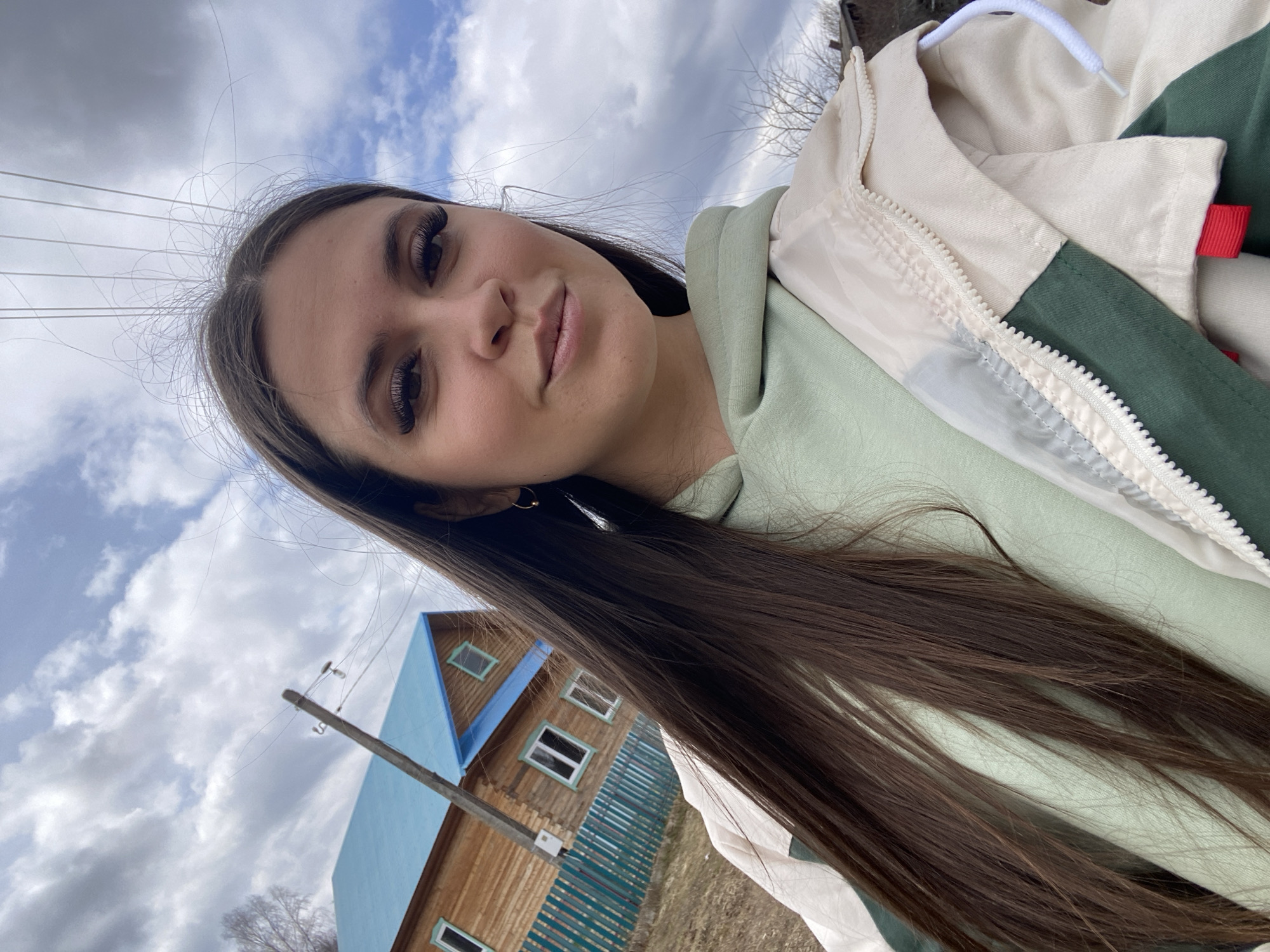 A selfie photograph taken outdoors in Zhigalki, Russia. The subject is a young woman with long, dark brown hair wearing a light green hoodie under a cream and green jacket. Her hair partially obscures a wooden house with a blue roof and white-framed windows. A utility pole with wires is also visible in the background. The sky is partly cloudy.