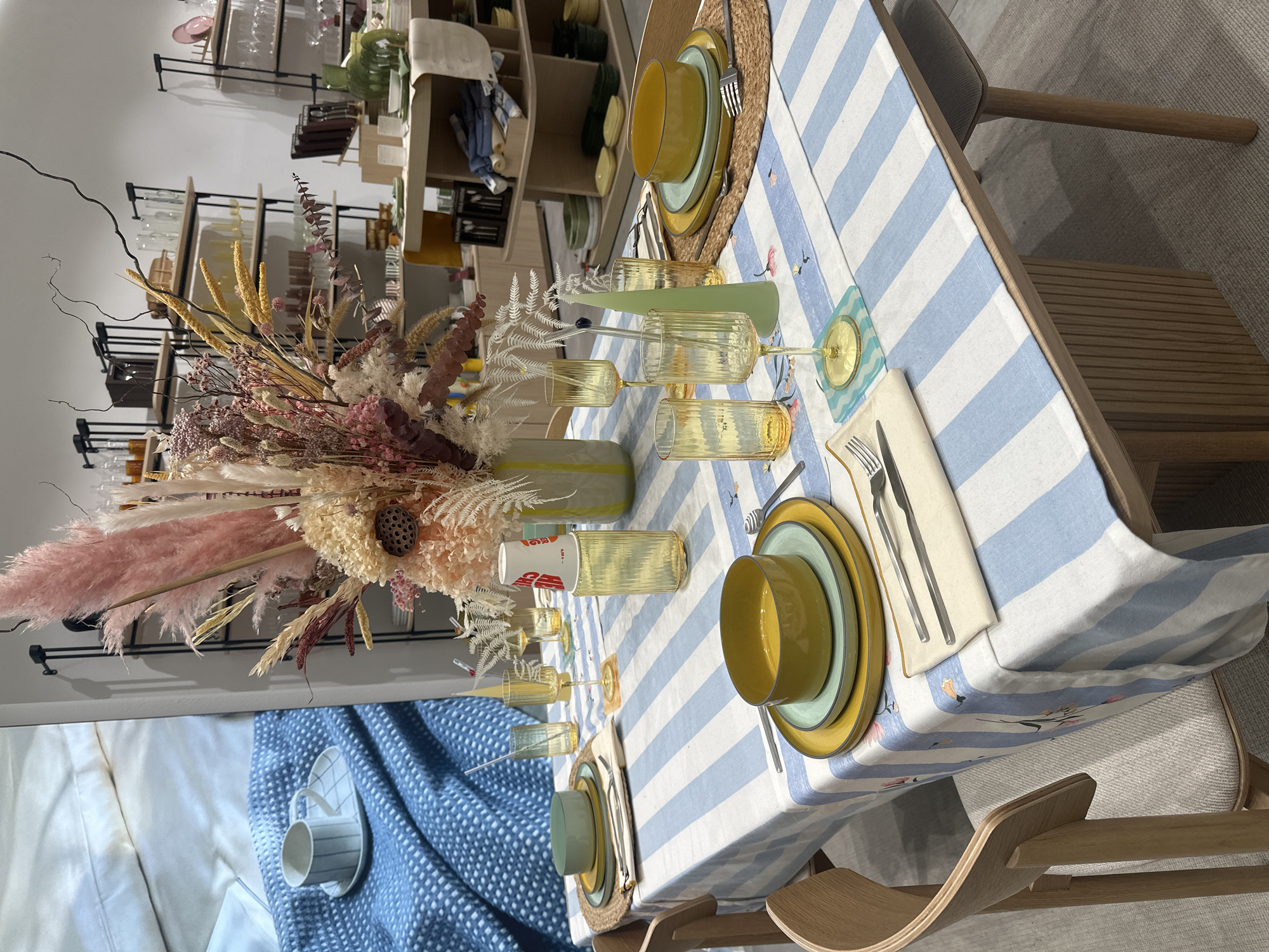 The image depicts a table setting inside a store in Родниковское сельское поселение, Ukraine. The table is covered with a blue and white striped tablecloth, and place settings for two people are visible. Each setting includes a yellow bowl, a green plate, a yellow plate, silverware, and a napkin. Woven placemats sit beneath the plates. Two yellow-tinted glasses are set on either side of a yellow-striped vase, which holds a large, dried flower arrangement. In the background, shelving units display various housewares and decorative items. A couch or bed is partially visible on the left side of the frame. Chairs are situated on opposite sides of the table.