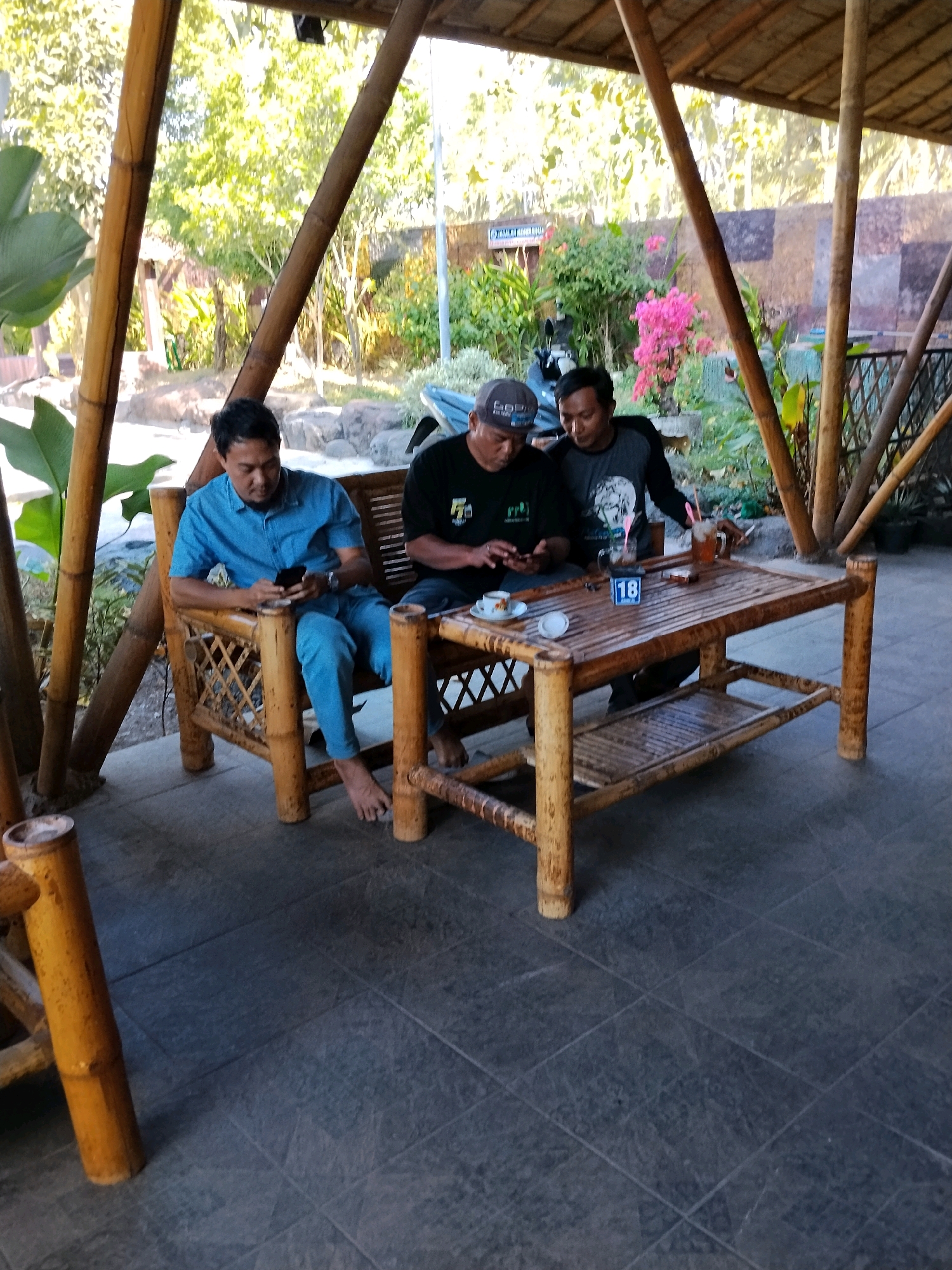 Three men are seated at a bamboo table under a bamboo roof. The man on the left, wearing a blue shirt and blue pants, is looking at his phone. The man in the middle, wearing a black t-shirt and a grey cap, is also looking at his phone. The man on the right, wearing a grey t-shirt with a design, is also looking at his phone. The table has two cups of drinks, a saucer with a small cup, a cigarette, and a number 18 displayed. The environment appears to be an outdoor seating area with bamboo furniture and lush greenery in the background, suggesting a restaurant or cafe setting. The overall lighting suggests it is daytime.
