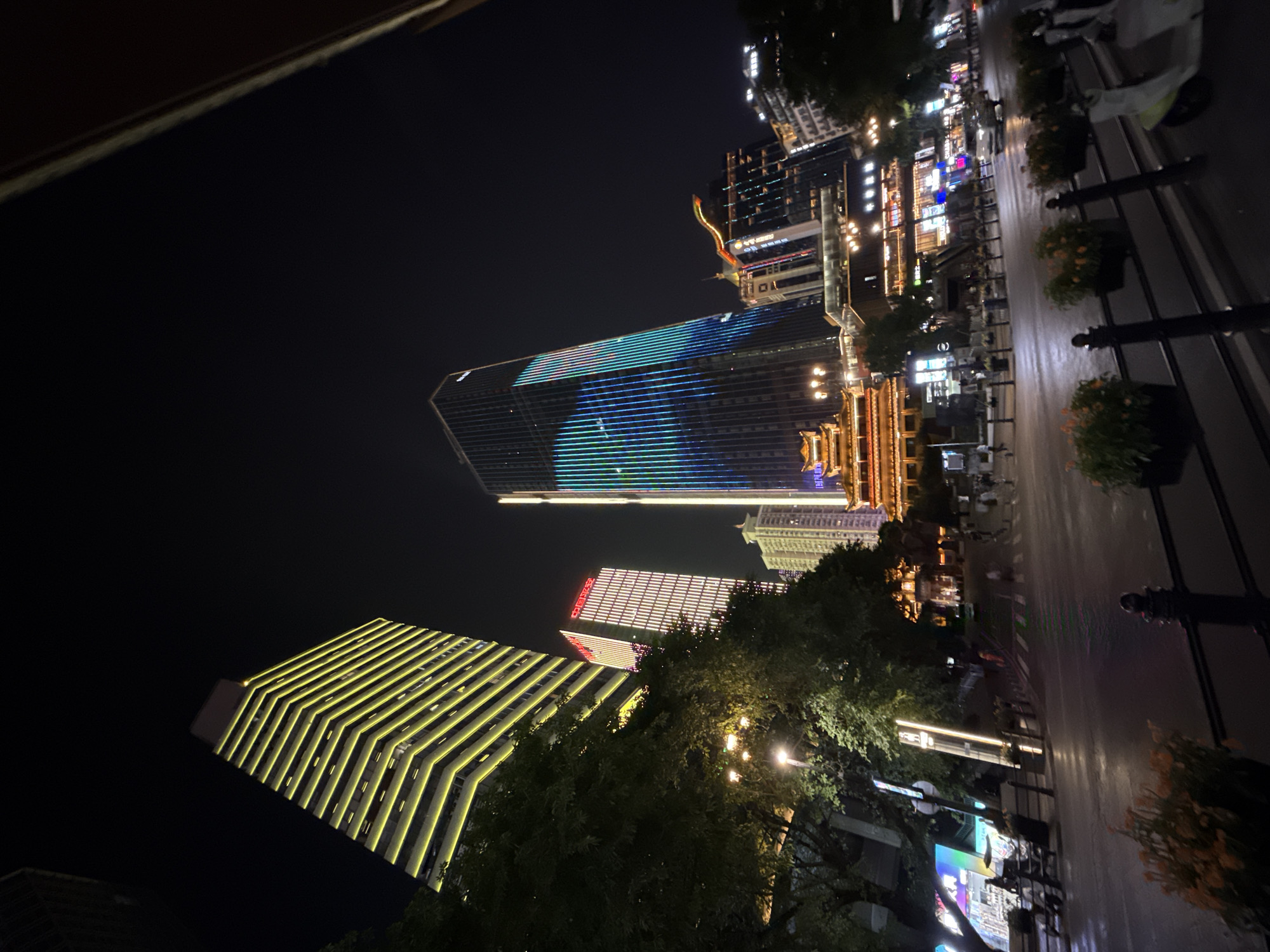 The image shows a nighttime street scene in the Yuzhong District of China, featuring modern architecture and vibrant city lights. In the foreground, a wet asphalt road reflects the surrounding illumination. Several dark metal railings with decorative posts line the edge of the sidewalk, interspersed with planters containing orange and green foliage. A few scooters are parked along the sidewalk.
Dominating the background are several tall buildings. The leftmost building is a skyscraper with a distinctive zigzag pattern of yellow lights running vertically along its facade. To its right, another building features a pattern of white lights. Further to the right, a prominent skyscraper displays a dynamic light show with vertical stripes of blue and green. In front of these modern structures, a building with traditional Chinese architectural elements, including a tiered, pagoda-like roof, is illuminated with warm lights. Other buildings with illuminated windows and signage are also visible, contributing to the bustling urban atmosphere.
People are sparsely scattered throughout the scene, appearing as blurred figures walking on the sidewalks or crossing the street. A white car is seen driving on the road, its headlights cutting through the darkness. The sky is a deep, dark black, typical of nighttime in a city with significant light pollution. The overall impression is one of a modern, energetic urban environment at night.