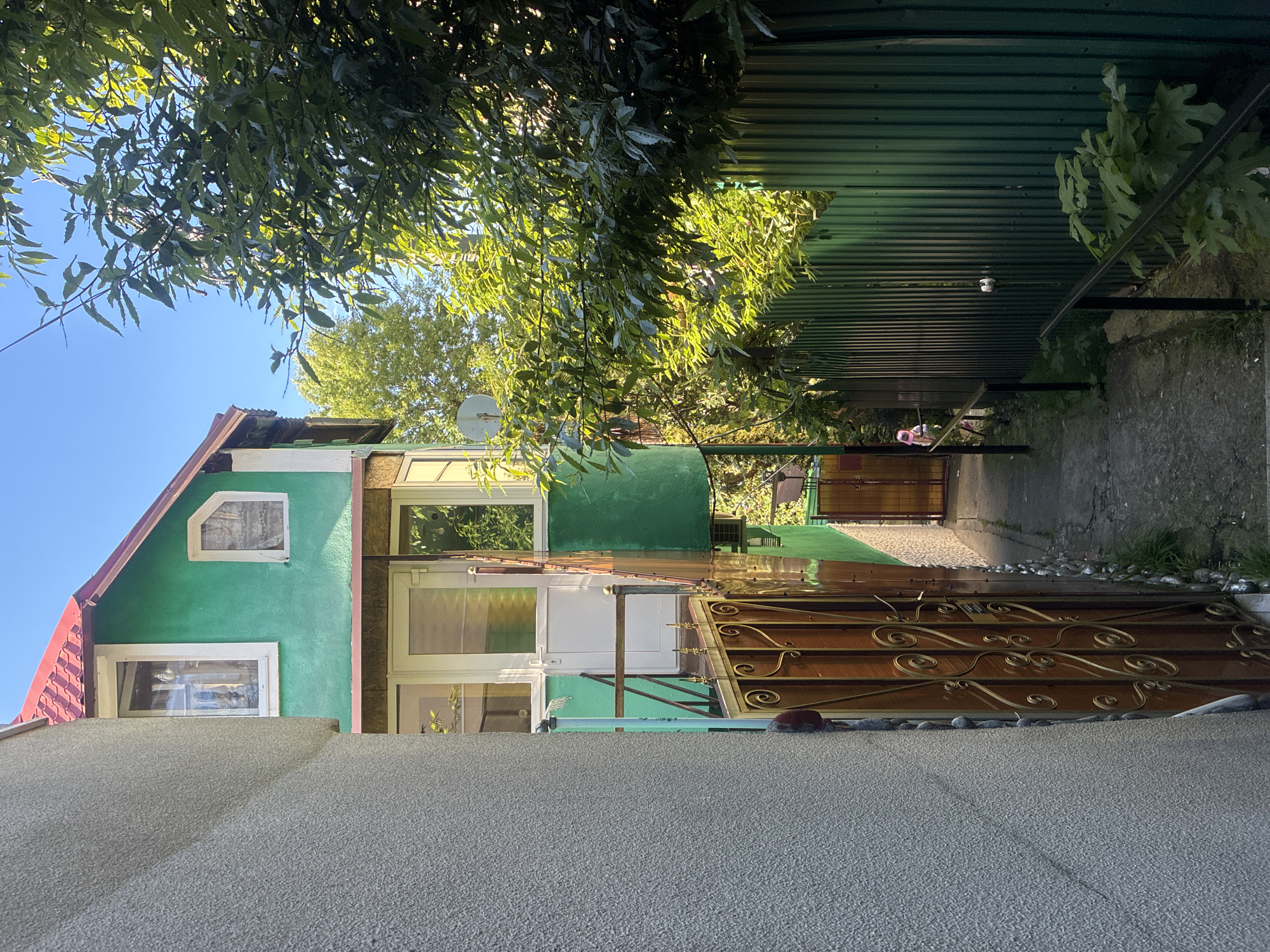 This image captures a vibrant, sunlit outdoor scene, likely in a residential area of Sochi, Russia. The perspective is looking down a narrow pathway or alleyway situated between a house and a textured wall in the foreground.
On the left side of the frame, a house with a striking green exterior wall and a reddish-brown tiled roof is visible. It features several white-framed windows, including one with a unique angled top on the upper floor. A white satellite dish is mounted on the side of the house. In the immediate foreground, a light grey, textured wall or structure occupies the bottom portion of the image.
To the right of the house, a concrete path recedes into the distance, partially covered by an overhead canopy made of green corrugated metal. Sunlight filters brightly through the leaves of overhead trees and gaps in the canopy, creating a dappled effect on the path. A prominent feature along this path is a decorative dark wooden gate adorned with ornate golden metalwork, with small stones lining its base. Further down the path, under the canopy, a light bulb fixture is visible.
Lush green tree foliage fills the top of the frame, with bright sunlight illuminating the leaves against a clear blue sky. The overall impression is one of a warm, sunny day, likely late morning or early afternoon, given the strength and quality of the light. No people are visible, and no text can be discerned in the image. The scene conveys a sense of peaceful domesticity.