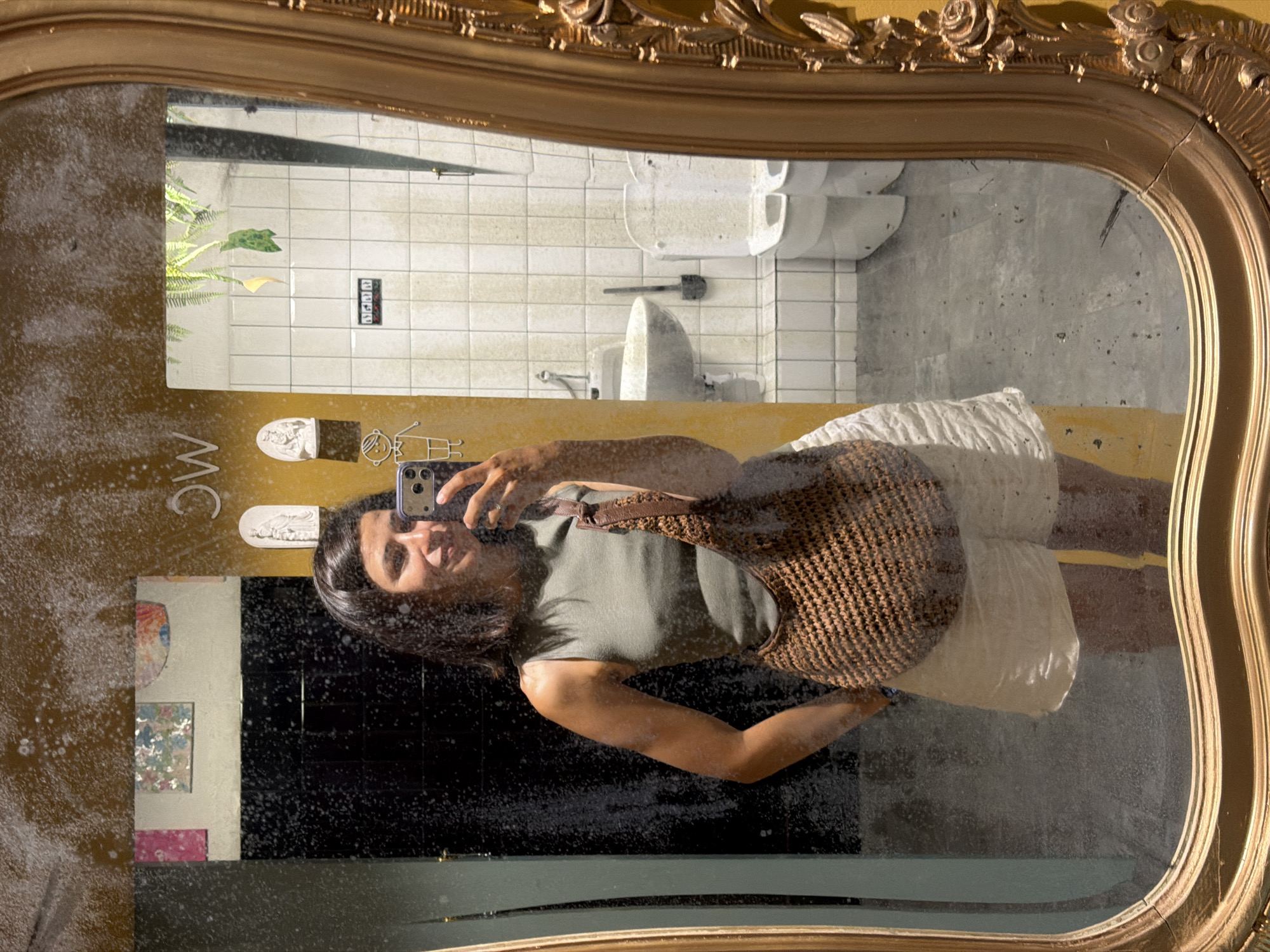 A young woman with dark hair takes a mirror selfie in a public restroom. She is wearing a sleeveless olive green top and white shorts, and is carrying a woven brown shoulder bag. Her face is partially obscured by her phone, which she holds up to take the picture. The restroom appears to be dimly lit, with white tiled walls and a grey concrete floor. A sink and two toilets are visible in the background. The mirror frame is ornate and gold-colored, with floral embellishments. The overall impression is of a casual, candid moment captured in a somewhat gritty, urban setting, potentially in a bar or club in Valencia, Spain, given the aesthetic.