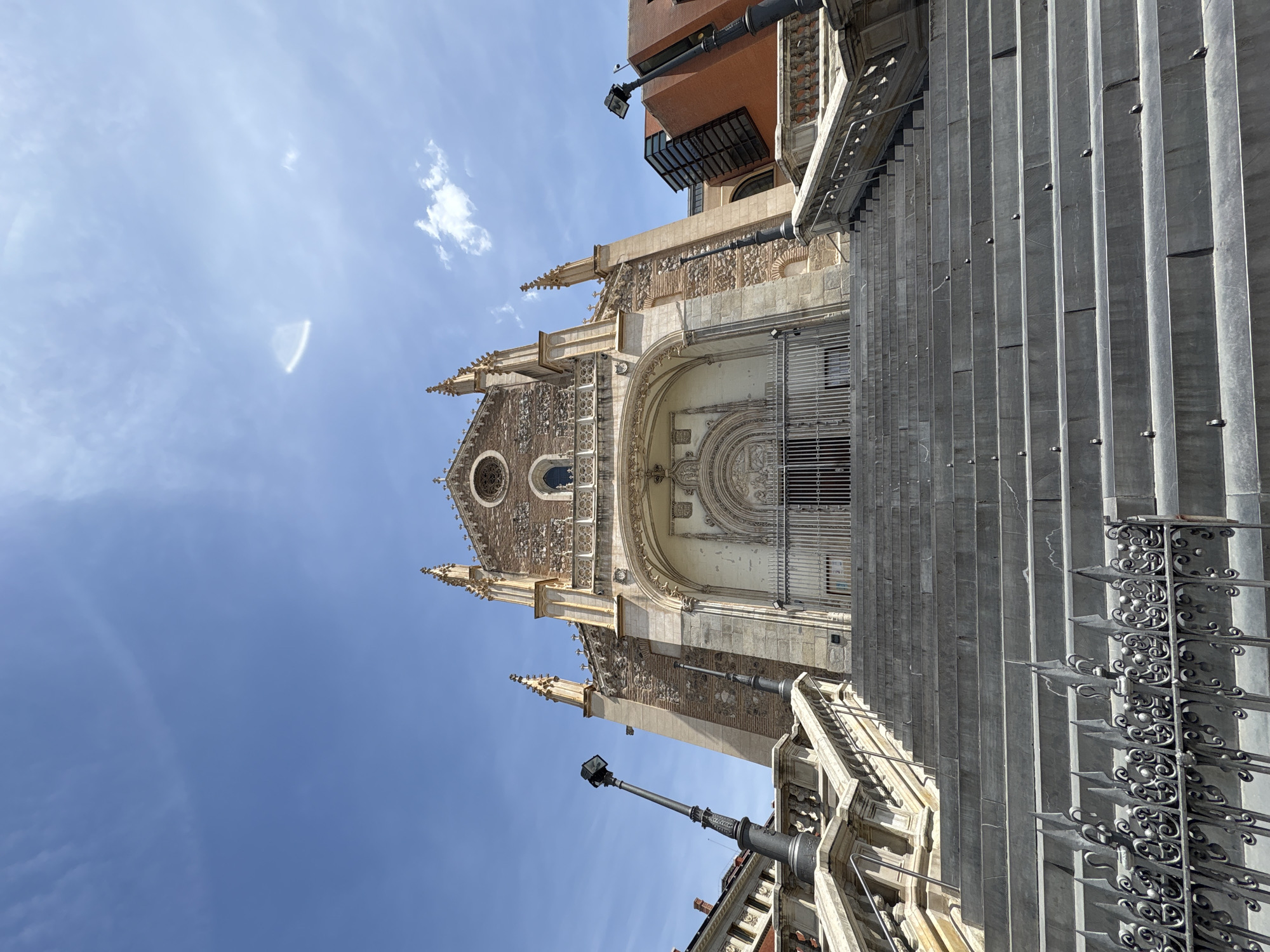 The image shows the Church of San Jeronimo el Real in Madrid, Spain. The building is constructed of light-colored stone, with intricate architectural details. The facade is visible, including the main entrance, rose window, and various spires and decorative elements. The church is partially obscured by a large set of stone steps, which lead up to the entrance. Metal railings run alongside the stairs. In the background, there is a section of a modern building, suggesting the church is located in a busy urban area. The sky above is blue with scattered clouds. There are lamp posts along the side of the building.