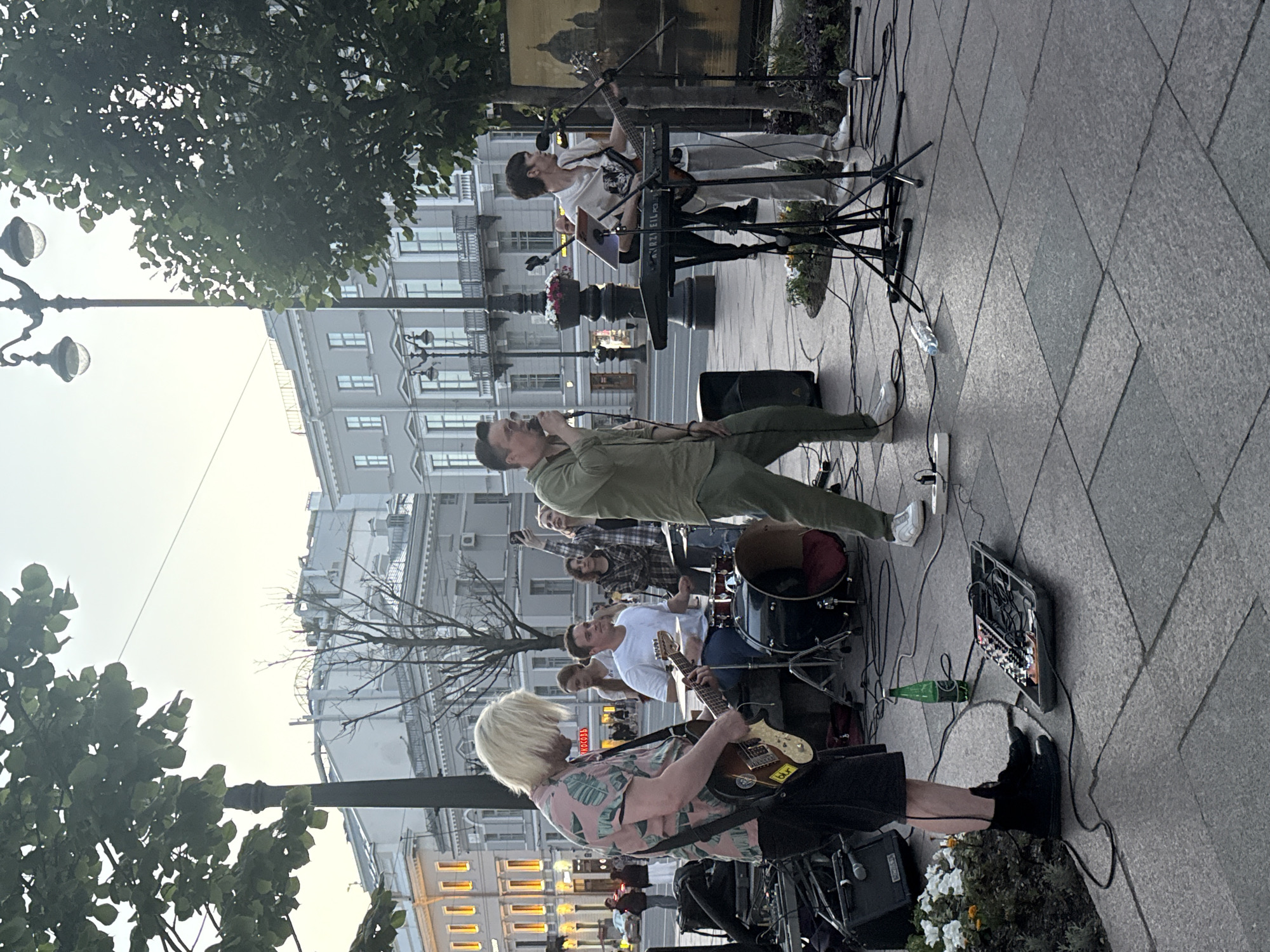 This media file shows a street performance by a band in Saint Petersburg, Russia. The band consists of at least four musicians. A singer in a green shirt is holding a microphone and appears to be the lead vocalist. Next to him, a guitarist with blonde hair in a pink floral shirt is playing a guitar. Behind them, a drummer is playing a drum set, and another person is playing a keyboard and guitar simultaneously. The scene is set on a paved sidewalk with buildings in the background. The time of day appears to be late afternoon or early evening, as indicated by the soft lighting and the buildings' illuminated windows. The weather seems pleasant. The overall mood of the image is energetic and lively, capturing a moment of live music being performed for an audience, though the audience is not clearly visible. Visible text includes a sticker on the guitar that reads "BLUR" and another on a pedalboard that is illegible.