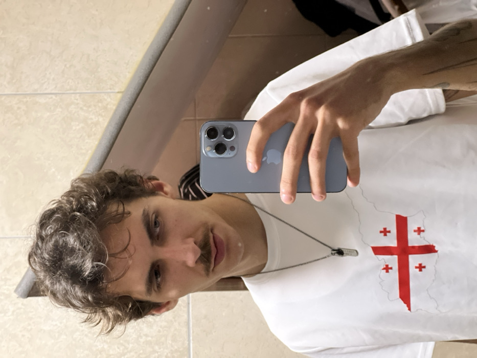An indoor mirror selfie depicts a young adult male with medium-length curly brown hair and a mustache. He wears a white short-sleeved t-shirt featuring a red St. George's cross over a map outline of Georgia, along with a silver-colored chain necklace with a dark rectangular pendant. A tattoo is partially visible on his right forearm. He holds a light blue/silver smartphone with a triple camera array in his right hand, angled downwards, reflecting the scene. The background consists of light-toned square tiled walls and a grey horizontal ledge. The setting is an interior space, likely a bathroom or changing room, located in Budva, Montenegro.