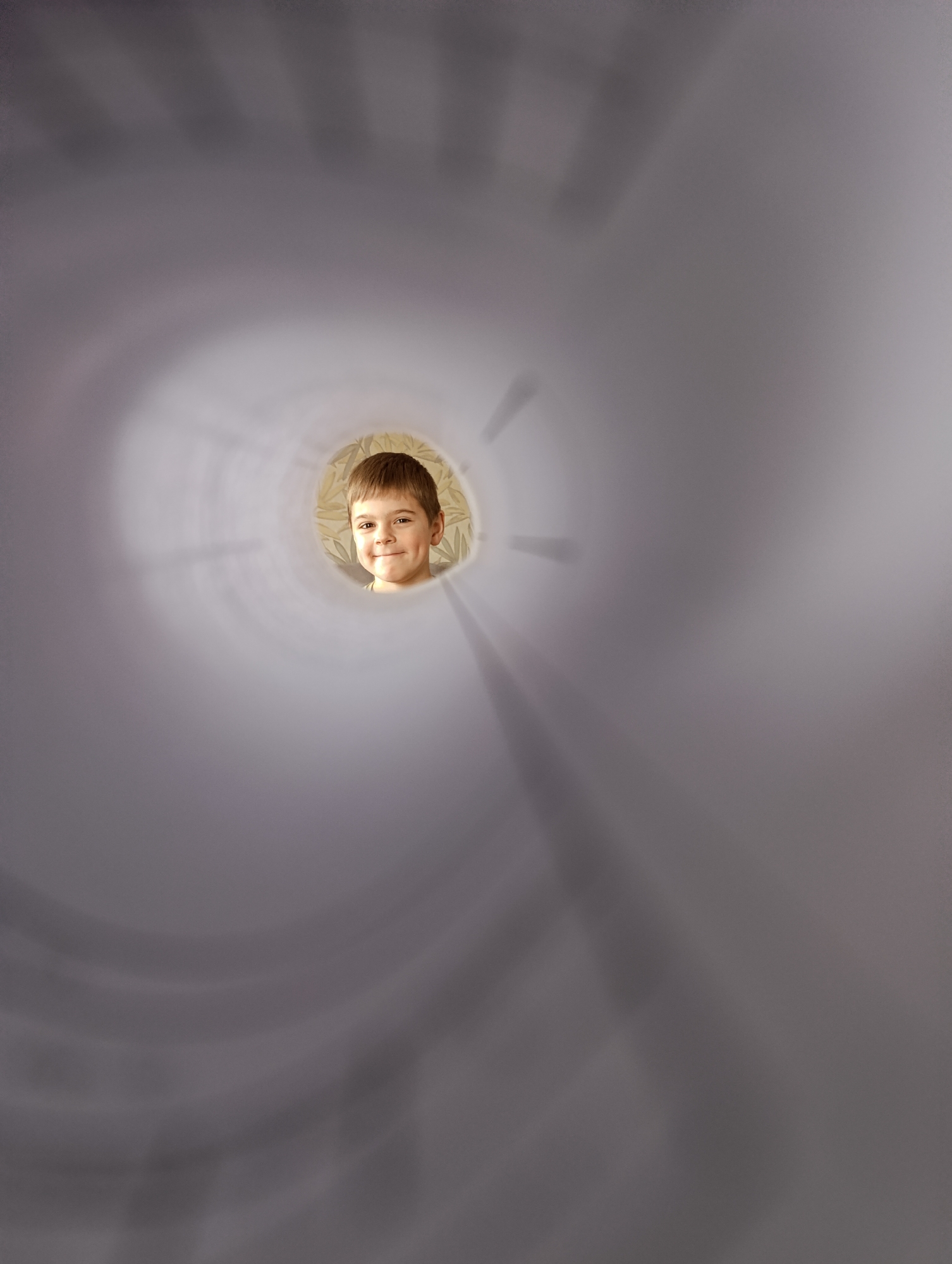 A child's face is visible through the center of a blurred, swirling, gray-toned object. The boy has short, brown hair, a small smile, and is centered within the circular opening. The swirling effect obscures any background details that would indicate a location. It is not possible to determine if the location is in Фоки, Russia.