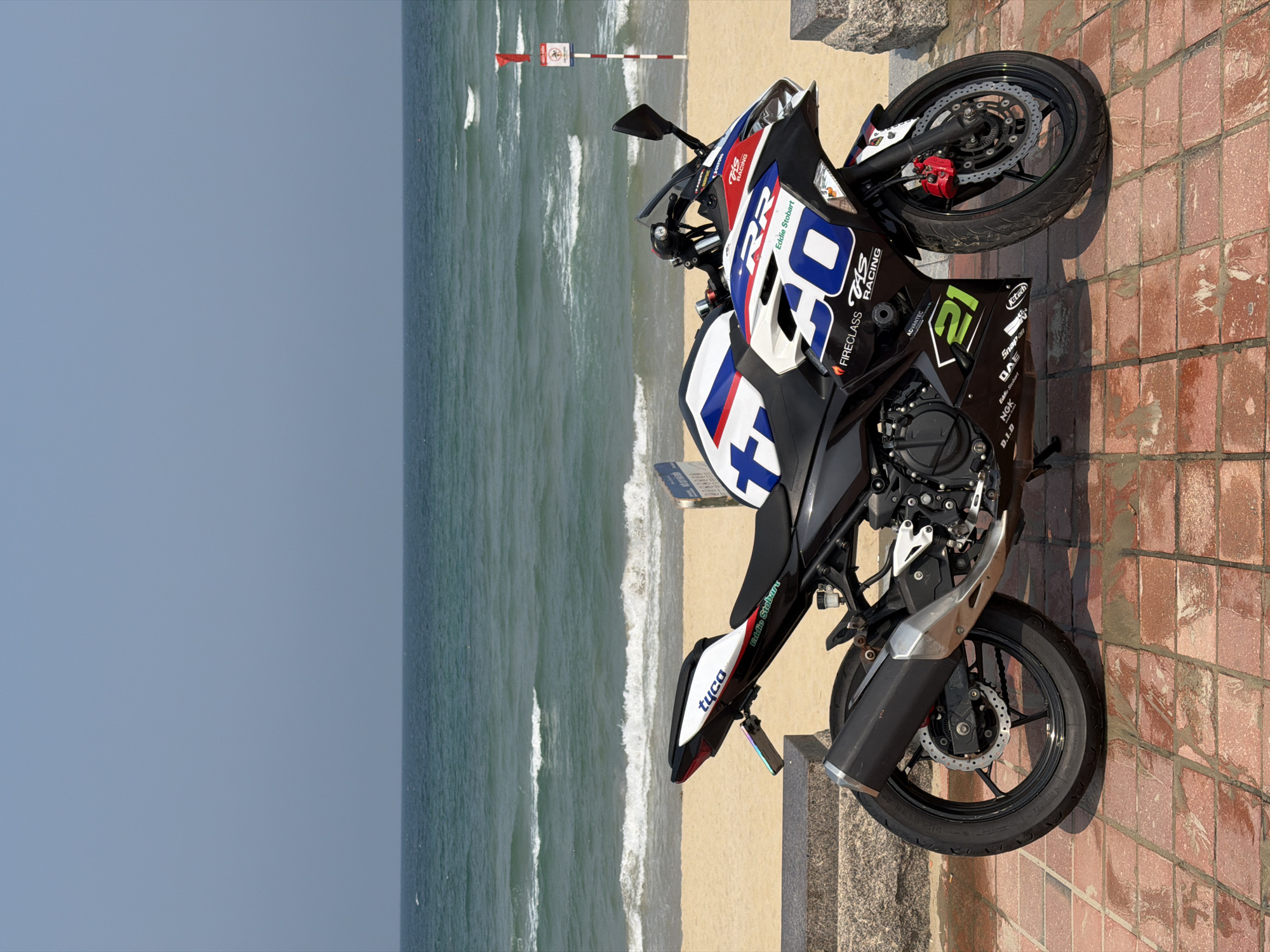 Here's a detailed description of the image:
**Scene:** An outdoor shot featuring a motorcycle parked on a brick path, with a scenic backdrop of the ocean and sky.
**Subject:** The primary subject is a black and white racing motorcycle with branding and decals, parked on a brick-paved area.
**Location:** The scene appears to be along a coast, likely in the city of Đà Nẵng, Vietnam. This is suggested by the beach, waves, and potential coastal landmarks.
**Environment:**
* **Sky:** The sky is a clear pale blue.
* **Ocean:** The ocean has visible waves rolling toward the shore.
* **Beach:** A sandy beach is visible between the water and the path.
* **Path/Promenade:** The motorcycle is parked on a brick path or promenade.
**Objects:**
* The motorcycle has various brand names and logos, like "Eddie Stobart" and "Tuco" printed on it.
* A signpost with a red flag and potentially a warning or restriction symbol is seen further down the beach.
* A curb or low wall separates the path from the beach.
* The background shows the ocean meeting the sky, creating a horizon line.
**Interactions/Actions:**
* The motorcycle is stationary and parked. There are no people visible interacting with it in the frame.
**Overall Impression:** The image captures a motorcycle parked by a beach, suggesting a moment of rest or display during a coastal ride. The details suggest a location in Đà Nẵng, Vietnam.