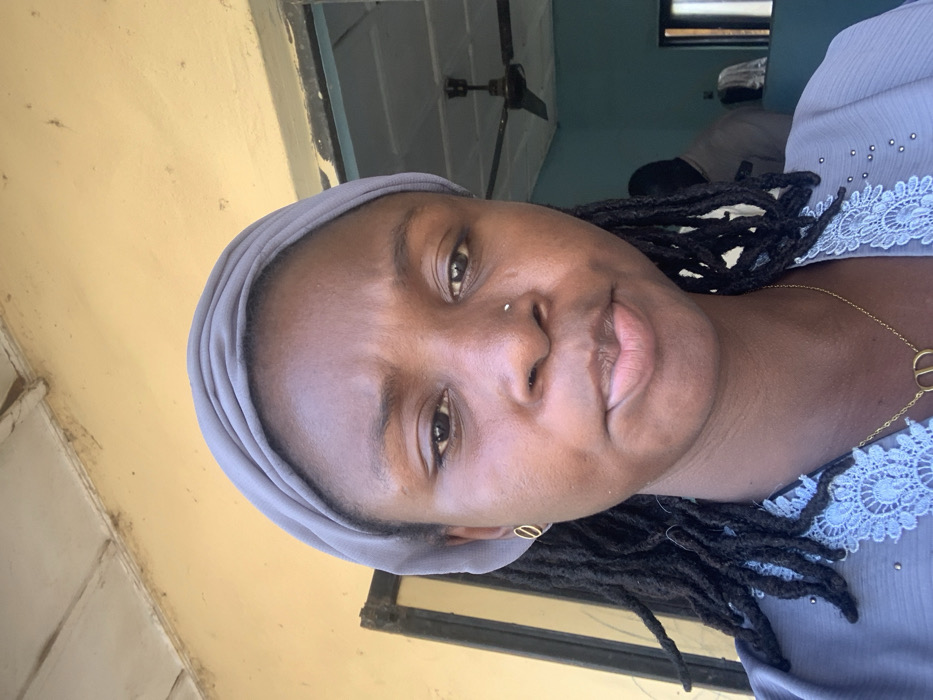 The image displays a close-up portrait of an adult female in Bojude, Nigeria, situated outdoors in front of a structure. The female has dark skin, dark dreadlocks partially covered by a light grey headscarf, and a small silver-colored stud piercing on her right nostril. She wears small gold-colored oval hoop earrings and a gold-colored chain necklace with a circular "CD" pendant. Her attire consists of a grey or light blue garment featuring light-colored lace trim. Her facial expression is neutral.
The background shows a faded yellow painted wall with visible discoloration and a wooden fascia or beam overhead. To the right, an open doorway reveals an interior room with light blue walls and a white ceiling. A dark-colored, stationary ceiling fan is suspended from the room's ceiling. The back of a partially visible individual, wearing a light-colored shirt, is present within this room. Another light-colored form is faintly visible deeper inside the room. The scene is illuminated by direct, bright light.