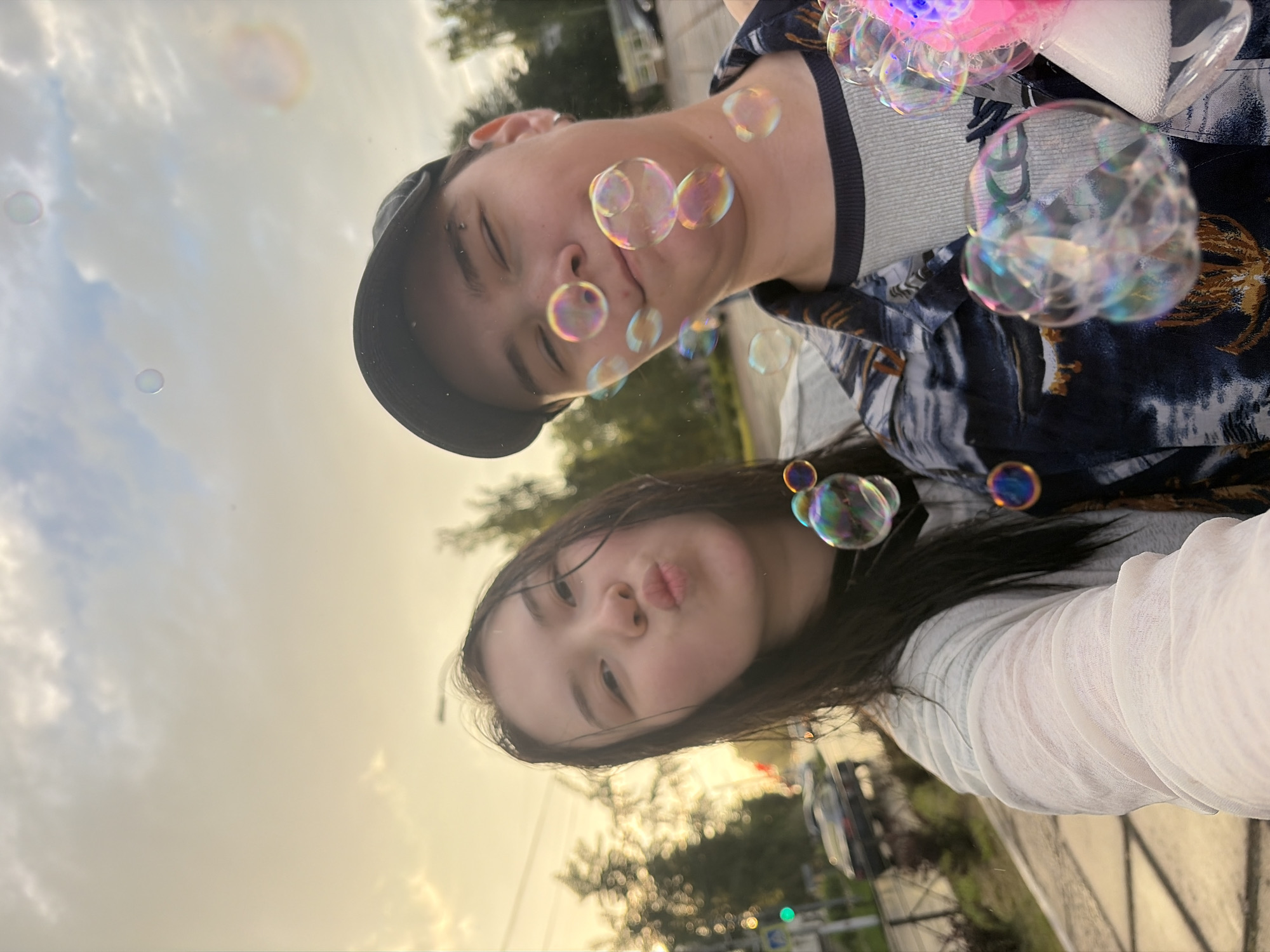 A selfie taken outdoors in Chaikovsky, Russia, features two young people and numerous soap bubbles. The background shows a street with a traffic light, cars, and trees, under a cloudy sky with golden sunlight breaking through.
The person on the left, a young woman, is positioned closer to the camera. She has long, dark hair that appears wet, and she is making a pouting face. She is wearing a light-colored, sheer top.
The person on the right, wearing a dark baseball cap and a patterned shirt over a grey undershirt, is looking towards the camera with their eyes closed and a slight smile. They have an eyebrow piercing and an earring.
Soap bubbles are scattered throughout the frame, some are clear, and others reflect the light with iridescent colors. One large bubble is prominently featured in the foreground on the right side of the image, near the person's shoulder. A portion of what appears to be a bubble machine, lit with pink and blue lights, is visible on the far right. The overall atmosphere suggests a casual, fun activity.