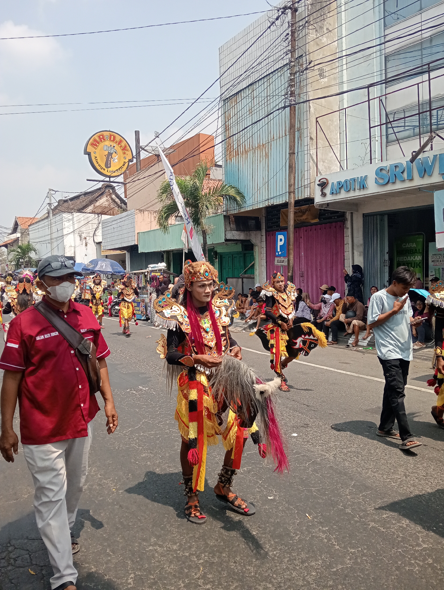 A daytime, eye-level outdoor medium shot shows a parade in progress on a street in Demak, Indonesia. The street is asphalt with some cracks. On the right side of the street, there are buildings with storefronts, one of which is labeled "APOTIK SRIWIJAYA". People are seated on the curb watching the parade. On the left side of the street, there are more buildings, and a yellow circular sign with a tool design is visible. The parade features performers in elaborate, traditional costumes. In the foreground, a man wearing a red shirt and a face mask is walking. Behind him, a performer dressed as a horse is walking, wearing a gold and red costume with a horse headpiece and flowing mane. Other performers in similar costumes are visible further back in the parade. The sky is blue with some clouds.