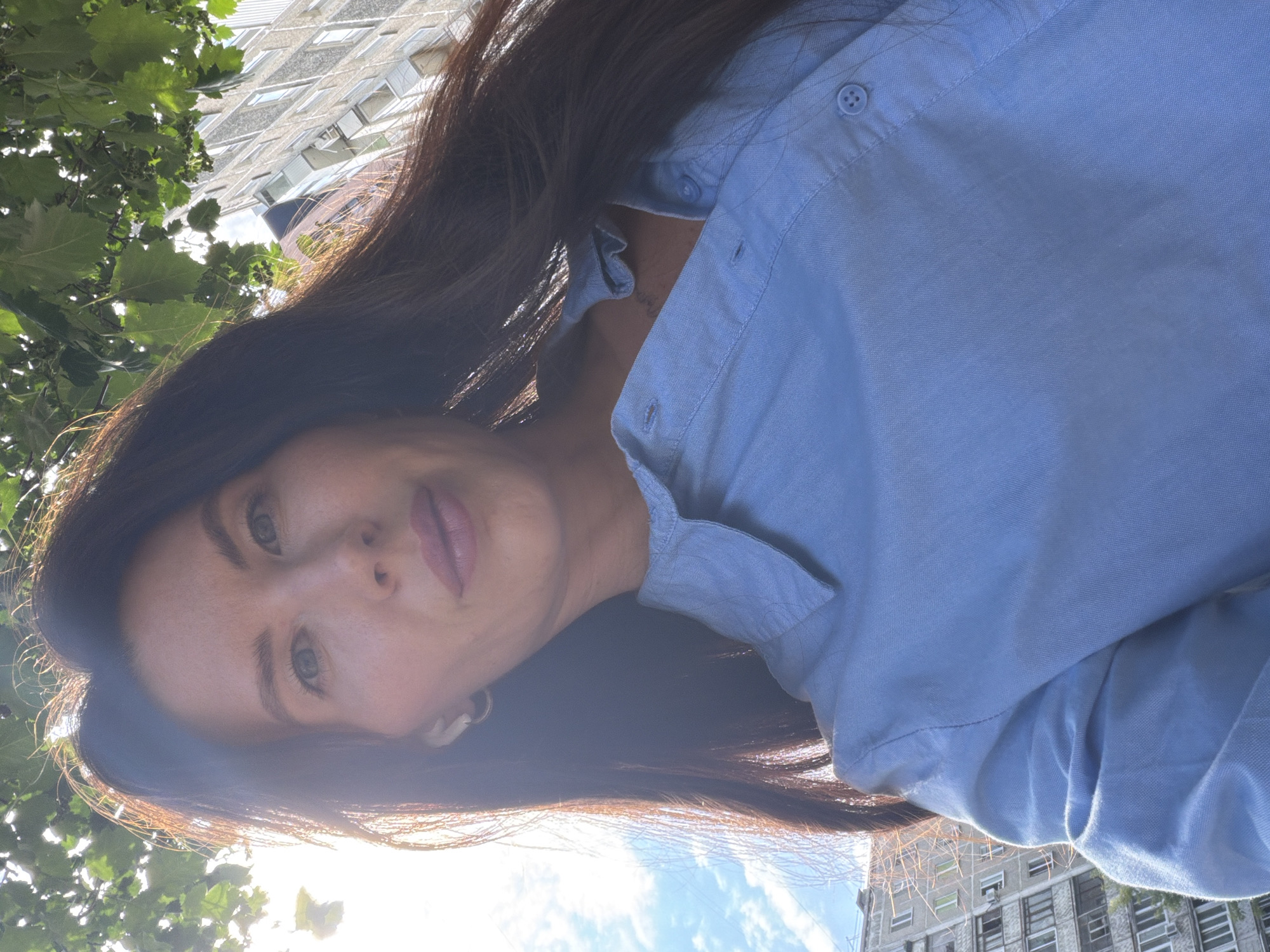 A low-angle, close-up selfie shows a woman outdoors in front of a building. She has long, dark brown hair, and is wearing a light blue collared shirt. Her facial features are visible, including her eyes, nose, and lips. She is also wearing small white earbuds and a gold hoop earring. The background includes green foliage from trees overhead, a bright sky with some clouds, and a multi-story residential building with many windows and balconies. The building appears to be a typical Soviet-era apartment block, common in cities like Perm, Russia. The lighting suggests it is daytime, possibly late afternoon or early morning due to the direction of the sunlight creating lens flare and illuminating the hair. The overall scene conveys an informal, personal moment captured outdoors.