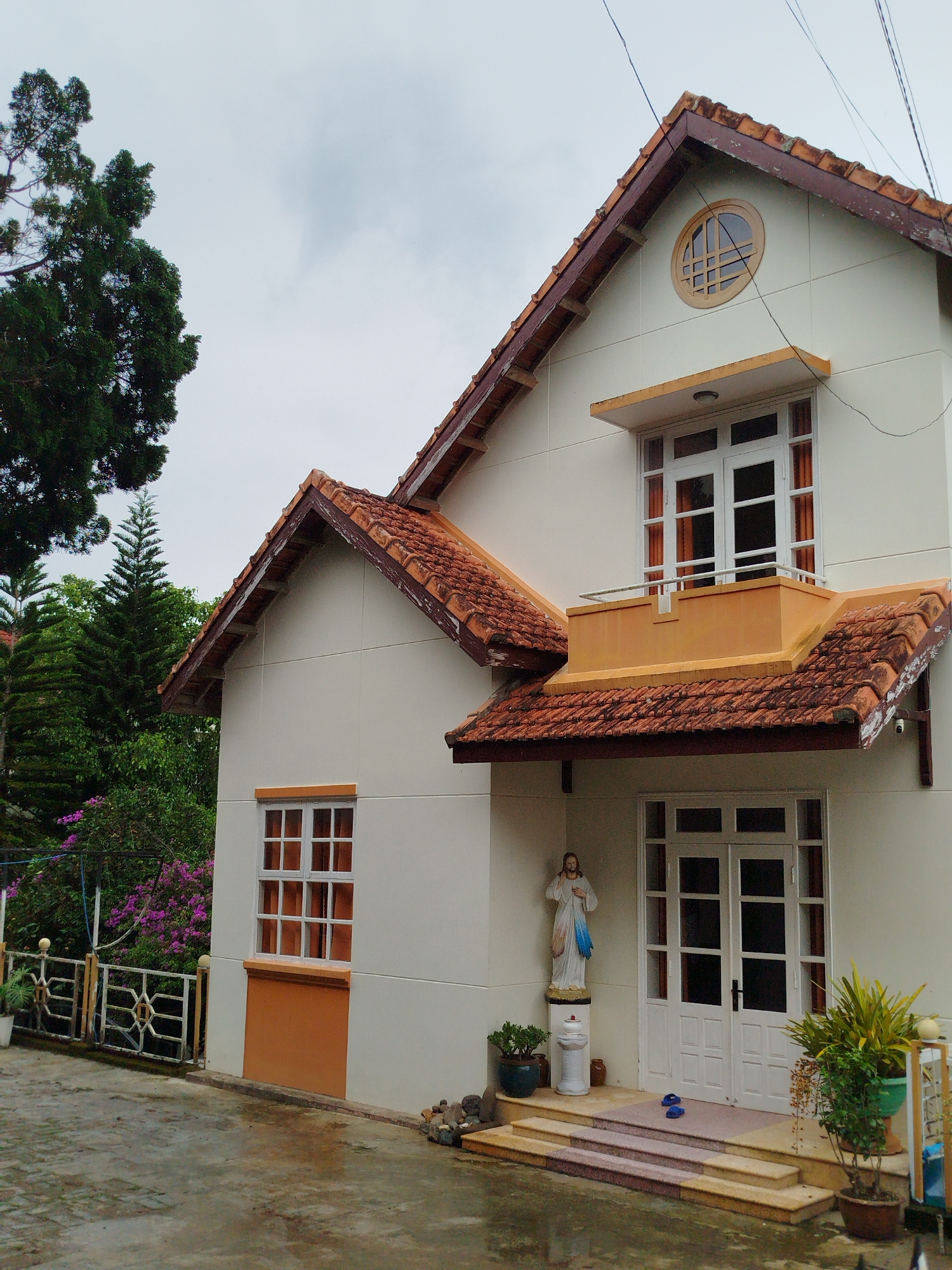 The image depicts a two-story house in Da Lat, Vietnam. The house is white with a terracotta tile roof. The roof has multiple gables and a decorative round window on the upper part of the front gable. There is a window with a balcony above the front door. The lower part of the house has a window on the side. The front door is white with a multi-pane design. A statue of Jesus Christ stands to the left of the door, next to a small fountain-like structure. Potted plants are placed on either side of the steps leading to the door. In front of the house is a wet pavement, indicating it might have rained recently. To the left, there's a fence bordering greenery, including trees and flowering plants. The sky is overcast. Power lines are visible above the house.
