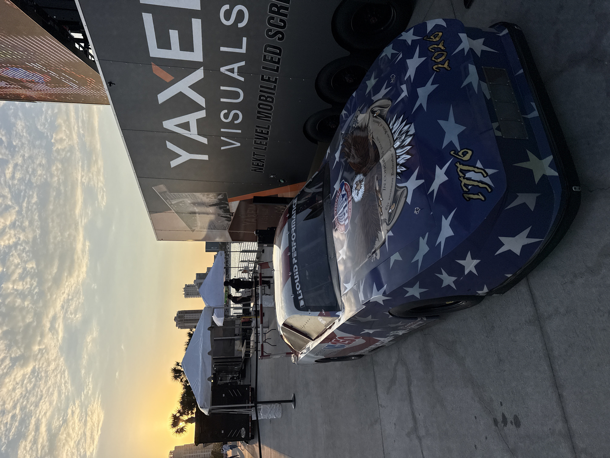 The image captures an outdoor event scene at dusk or dawn, situated on a concrete waterfront promenade in downtown Saint Petersburg, Florida, USA. A modified racing vehicle, likely a drag car, is prominently displayed in the foreground. Its body is adorned with an American flag-themed livery, featuring blue with white stars, a large bald eagle graphic on the hood clutching a scroll inscribed "IN CONGRESS JULY 4, 1776 The unanimous Declaration of the thirteen united States of America." The numbers "1776" and "2026" are painted in gold on the vehicle's rear, and "LIQUID PERFORMANCE" and "IHRA" branding are visible on its side.
To the right of the vehicle, a large black semi-trailer is parked, branded with white lettering "YAXEL VISUAL SOLUTIONS" and "NEXT LEVEL MOBILE LED SCREEN." A portion of a large LED display screen is visible above the trailer. In the background, several white canopy tents are erected along a waterfront railing, behind which a body of water is visible. Multiple individuals are present; two figures are discernible near the tents by the railing, one appearing to wear a dark uniform. Another individual walks in the far left background. The sky displays a warm, golden hue with scattered clouds, indicating sunrise or sunset. Distant city buildings and palm trees are silhouetted against the horizon, completing the urban waterfront setting.