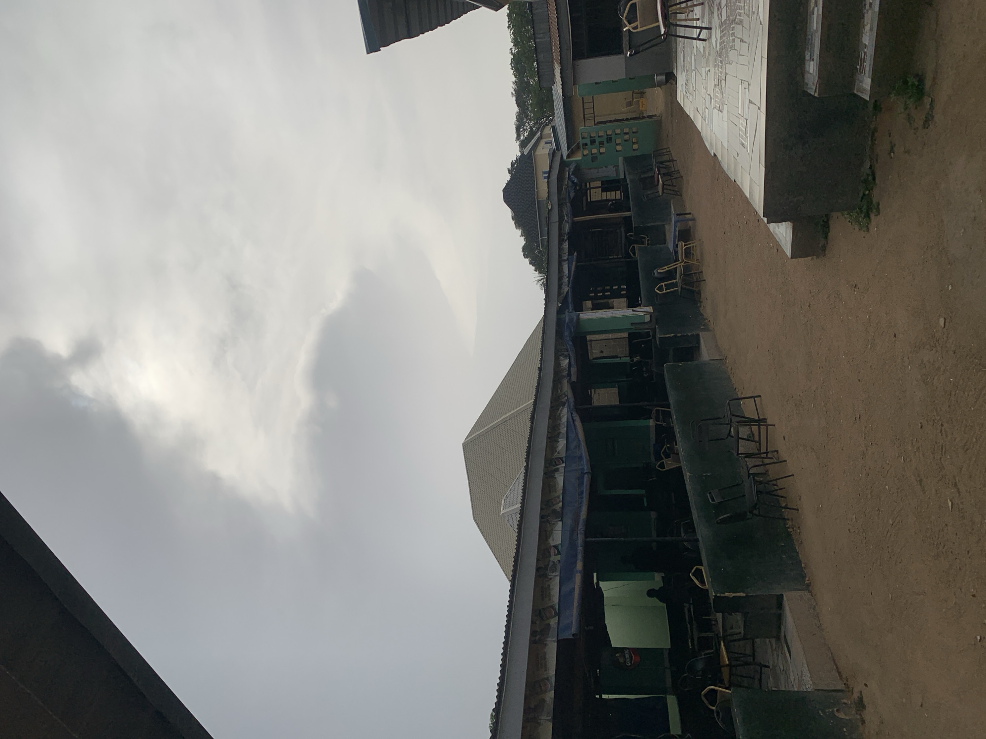 The scene depicts a row of dark green seating areas with metal chairs, likely an outdoor gathering space, situated in what appears to be New Stadium, Nigeria. The seating areas are positioned along a dirt ground, with a building featuring a pitched roof visible behind them. The sky is overcast. There is a person seated at a table in the shadows.