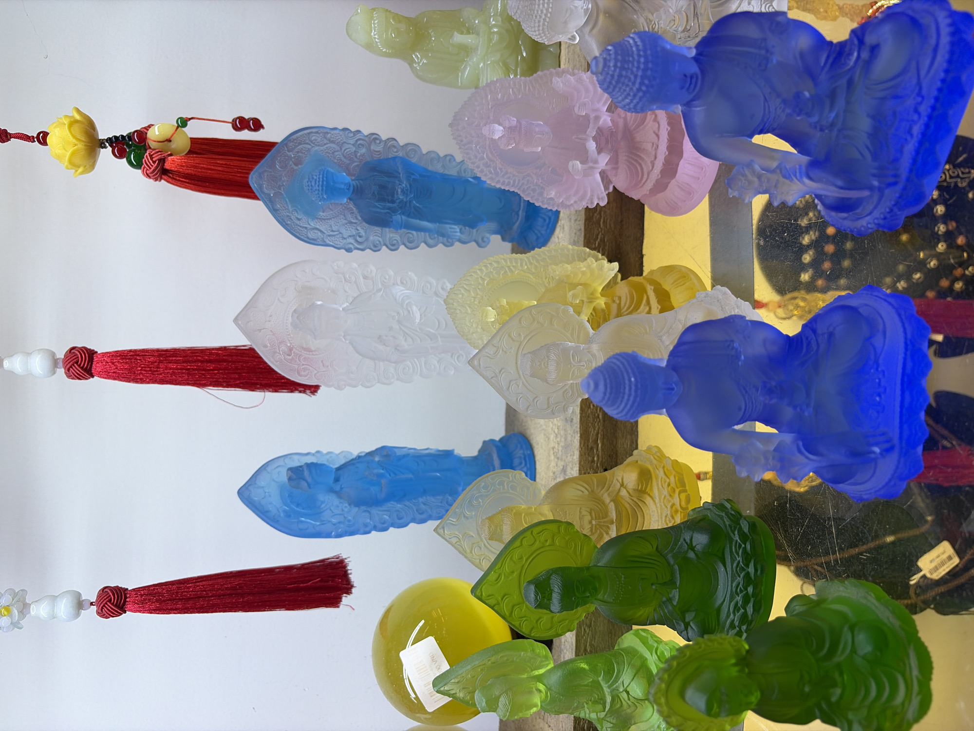 This image captures a display of various colorful translucent Buddhist figurines, likely made of glass or resin, arranged on shelves. The figurines are of different colors, including green, yellow, blue, pink, and clear. Some are standing, while others are seated in meditative poses. Behind the figurines, red tassels hang from decorative strings, possibly part of a larger hanging ornament. In the background, a plain white wall serves as the backdrop. There is no identifiable signage or architectural detail to pinpoint a specific location within Đà Nẵng, Vietnam, although such religious or decorative items are commonly sold in markets or souvenir shops in tourist areas of the city. The lighting appears to be bright, possibly from artificial sources, illuminating the translucent quality of the figurines and creating reflections on the surface they are displayed upon. There are no people visible in the image.