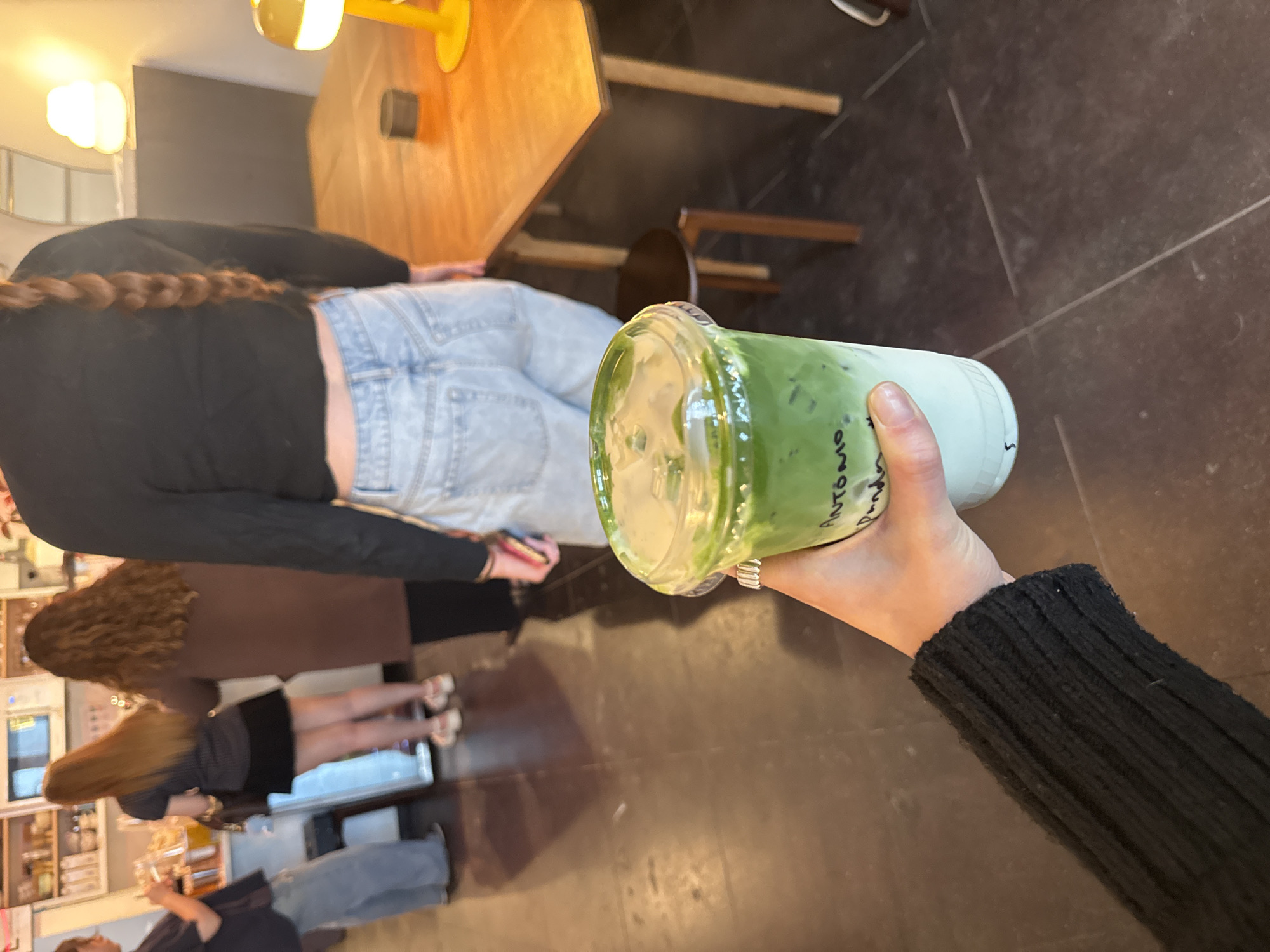 A hand in a black sweater sleeve holds a plastic cup filled with a green and white drink with writing on the side, positioned in front of a partial view of a restaurant interior. The restaurant features a light-colored wooden table and chairs, and multiple people are visible in the background. The photograph's perspective is from slightly above, looking downward. The location is Dallas, Texas, USA.