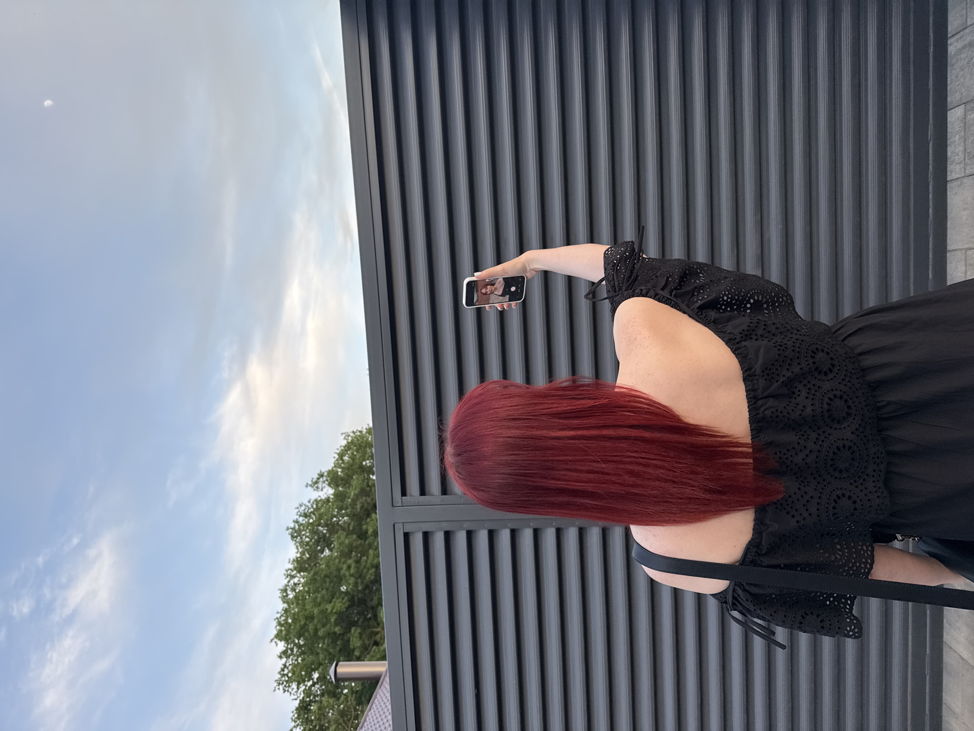 A woman stands outdoors, her back to the viewer, holding a smartphone up to take a photo or video. The phone's screen displays a person, presumably herself. The woman has shoulder-length, red hair and is wearing a black dress with eyelet detailing on the sleeves. She stands against a dark grey, corrugated metal wall. To the left, trees are visible in the background, and above, there is a pale blue sky with clouds. The location is a city of Unknown, Unknown.
