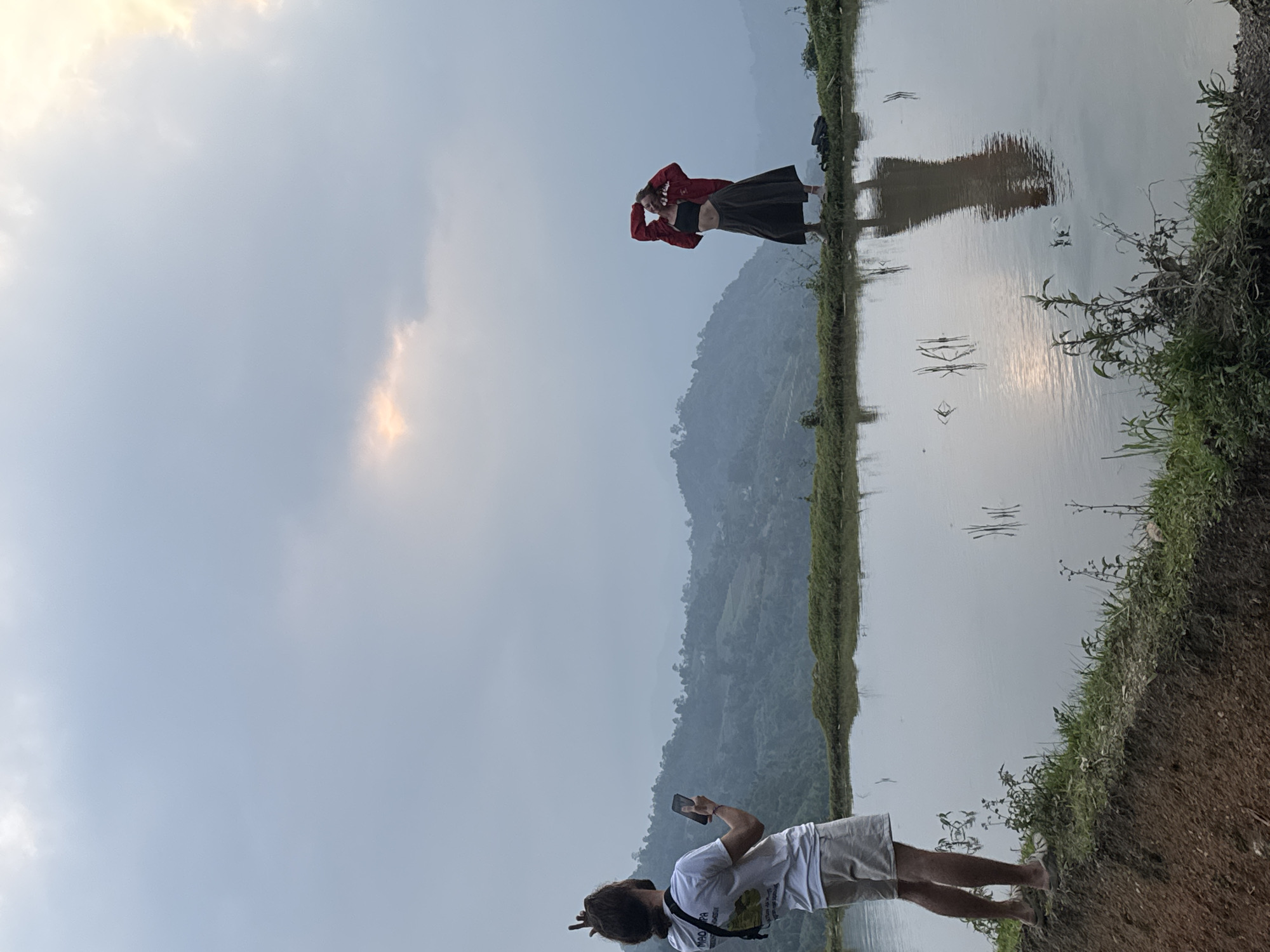 This image captures a scenic moment in a rural landscape, likely in Nậm Dịch Commune, Vietnam. The main subjects are two individuals interacting against a backdrop of water and rolling hills.
In the mid-ground, a person, possibly female, stands on a narrow bank beside a large body of calm water, which reflects the sky and the surrounding landscape. She is dressed in a red jacket over a black top and a dark skirt, with her arms raised, hands near her head, striking a pose. Her clear reflection is visible in the placid water. Behind her, a dark bag or backpack rests on the ground.
In the foreground, another individual, seen from behind, is holding a smartphone, seemingly capturing a photograph or video of the posing person. This person wears a white t-shirt with some faint text (possibly "MAO PA" or similar, and other unreadable words) and light-colored shorts. They have long, dark hair, possibly dreadlocks, tied up.
The setting features a soft, overcast sky with hints of warmer light, suggesting either early morning or late afternoon. Distant mountains or hills rise in the background, adding depth to the scene. The water surface is smooth, creating mirror-like reflections, indicating calm weather. The strip of land separating the subjects from the water appears earthy and lined with sparse vegetation. The overall atmosphere is tranquil, focusing on the human interaction within the expansive natural environment.