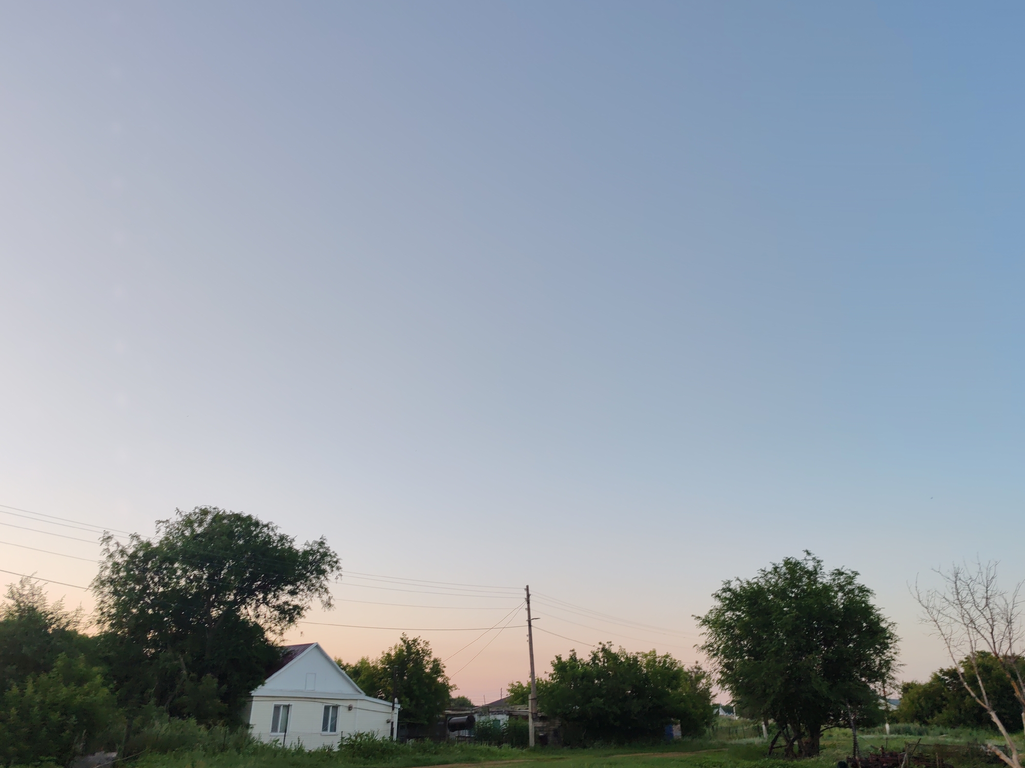 The image shows a rural scene with a clear sky at dusk. A small white house with a red roof is visible on the left side of the image, surrounded by lush green trees and vegetation. A telephone pole with wires runs across the scene. In the background, more trees and buildings can be seen. The overall impression is one of a quiet, serene evening in the countryside.